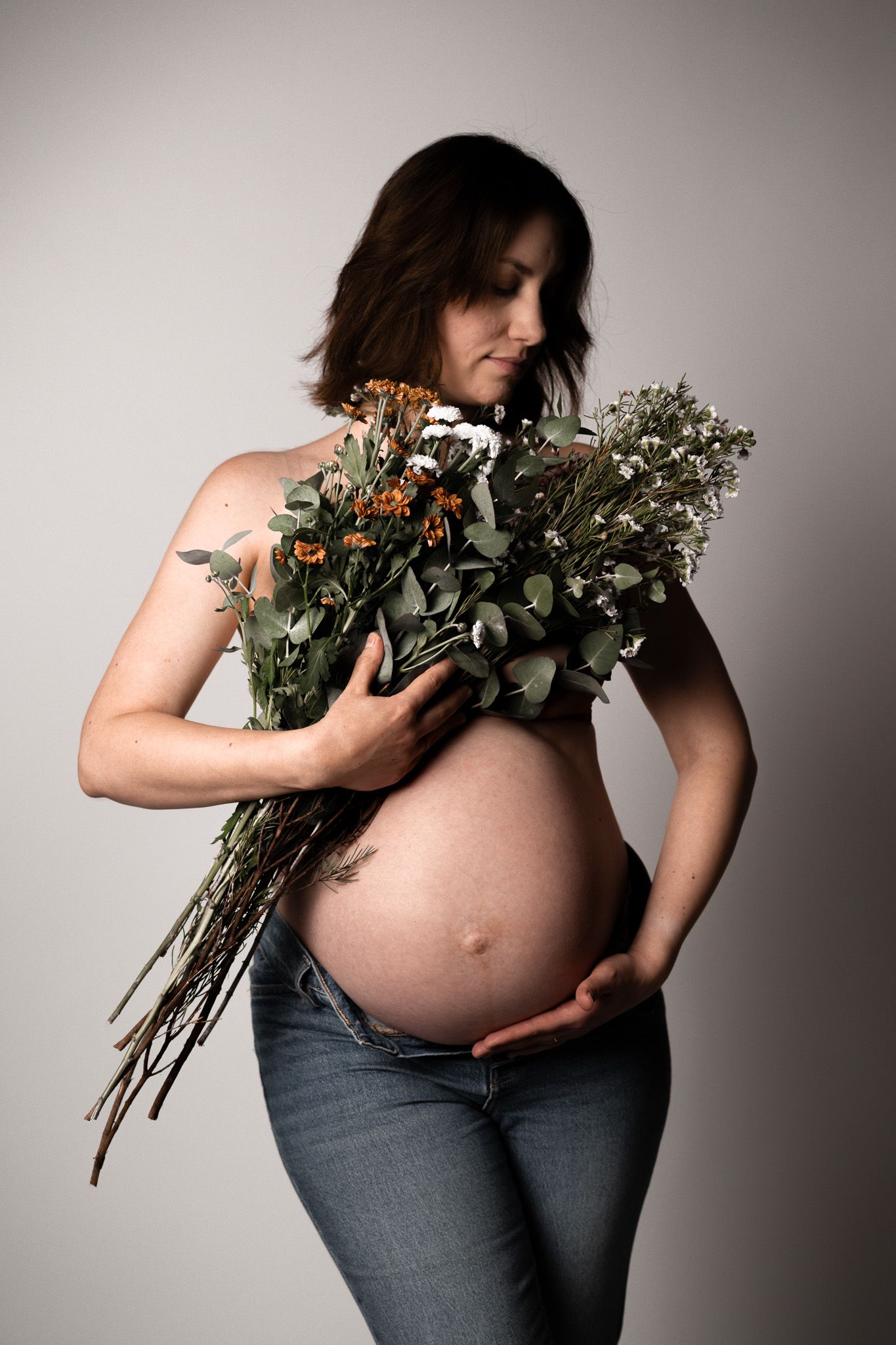 Eine junge Schwangere Frau die einen großen Blumenstrauss vor ihren großen Bauch hält und mit der anderen Hand ihren Bauch umrahmt.