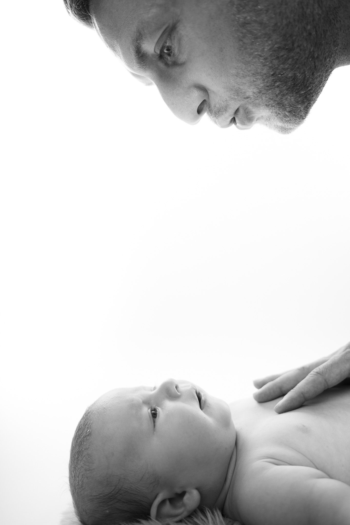 Ein Vater schaut liebevoll auf seinen neugeborenen Sohn nieder. Das Baby lächelt zurück. Schwarz-weiß Newborn- Fotografie