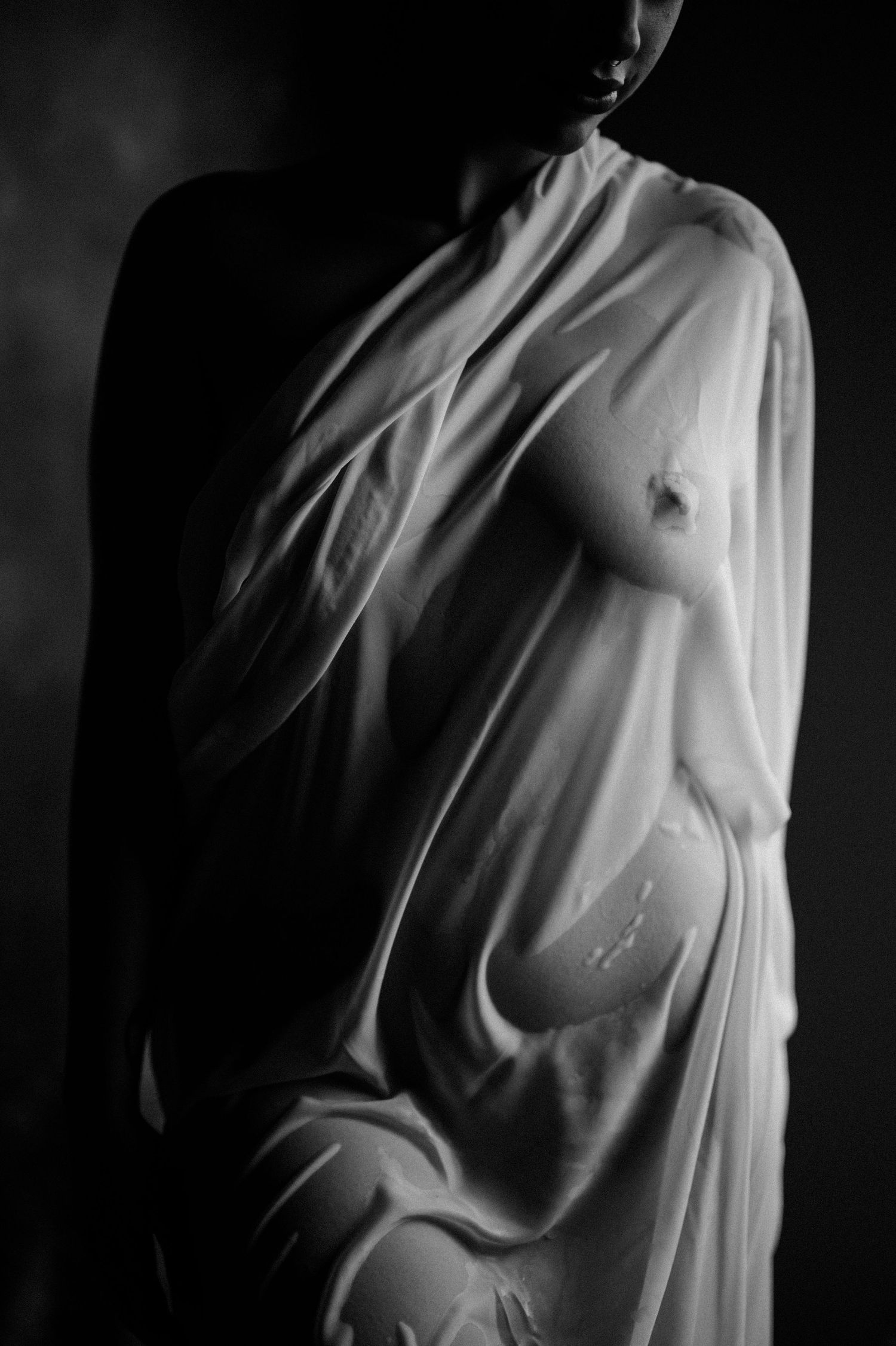 Black and white artistic boudoir photo of a woman draped in sheer white fabric against a dark background.