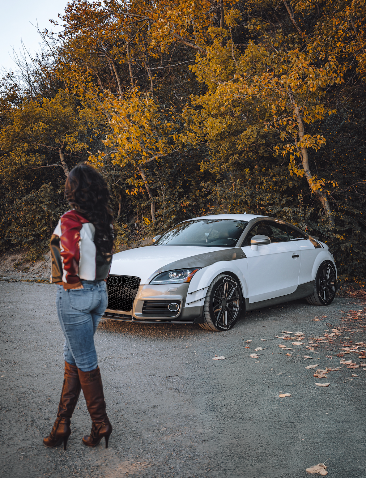 Audi TT parked under orange and yellow autumn tree with girl looking at the car