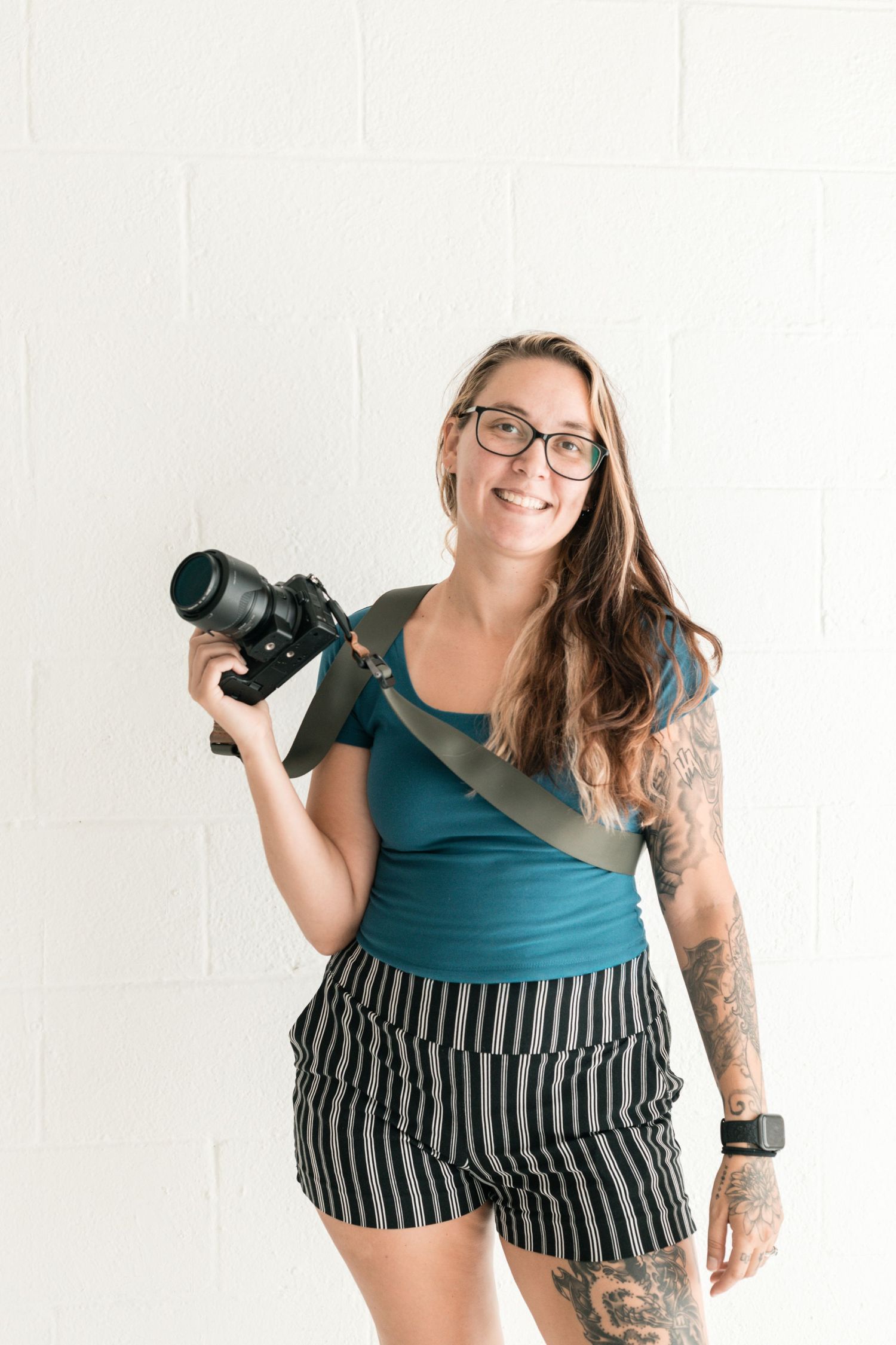 Photographer in teal top showing playful poses with camera in white studio setting.