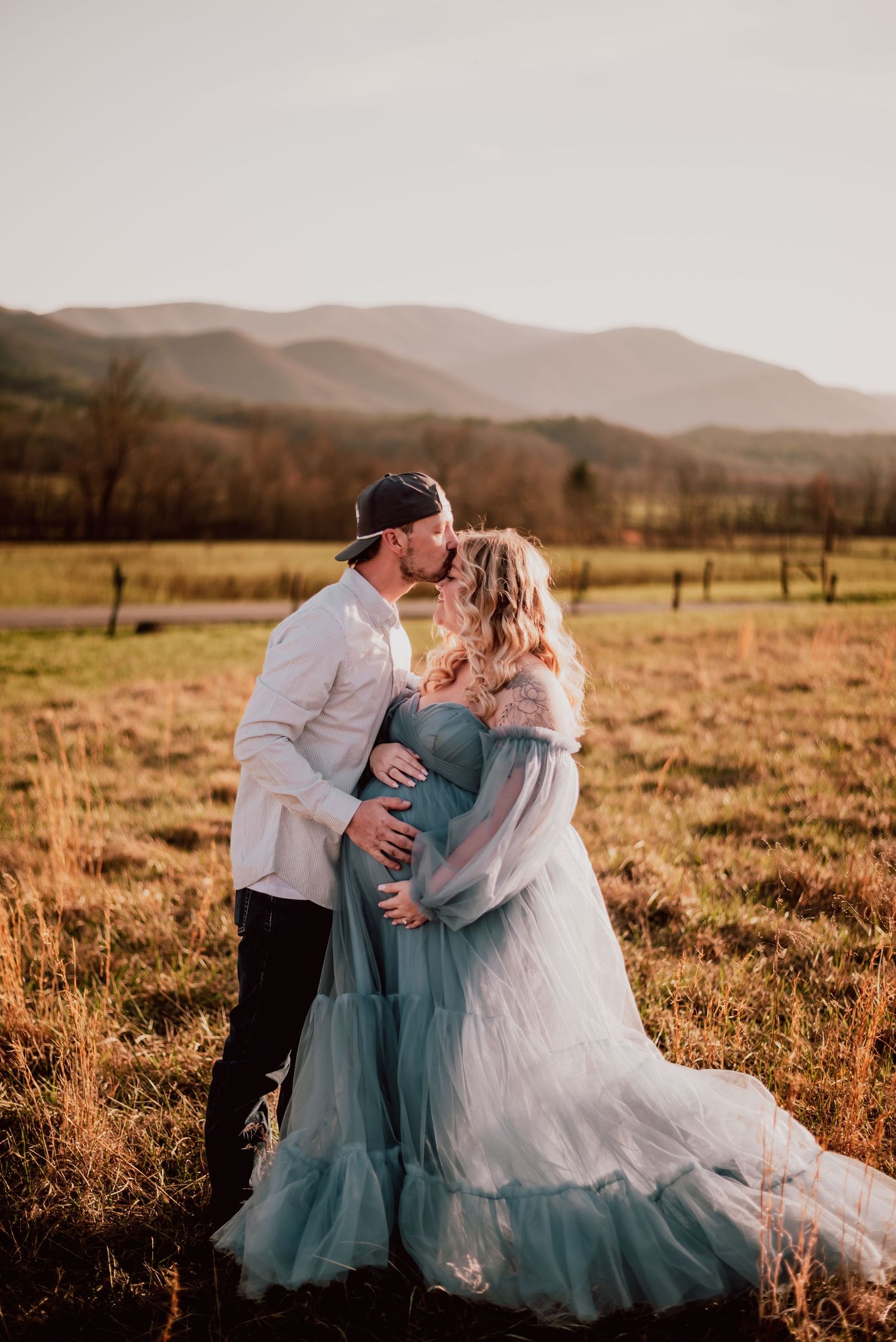 A romantic maternity photo of a couple embracing in a field at sunset with mountains in background.