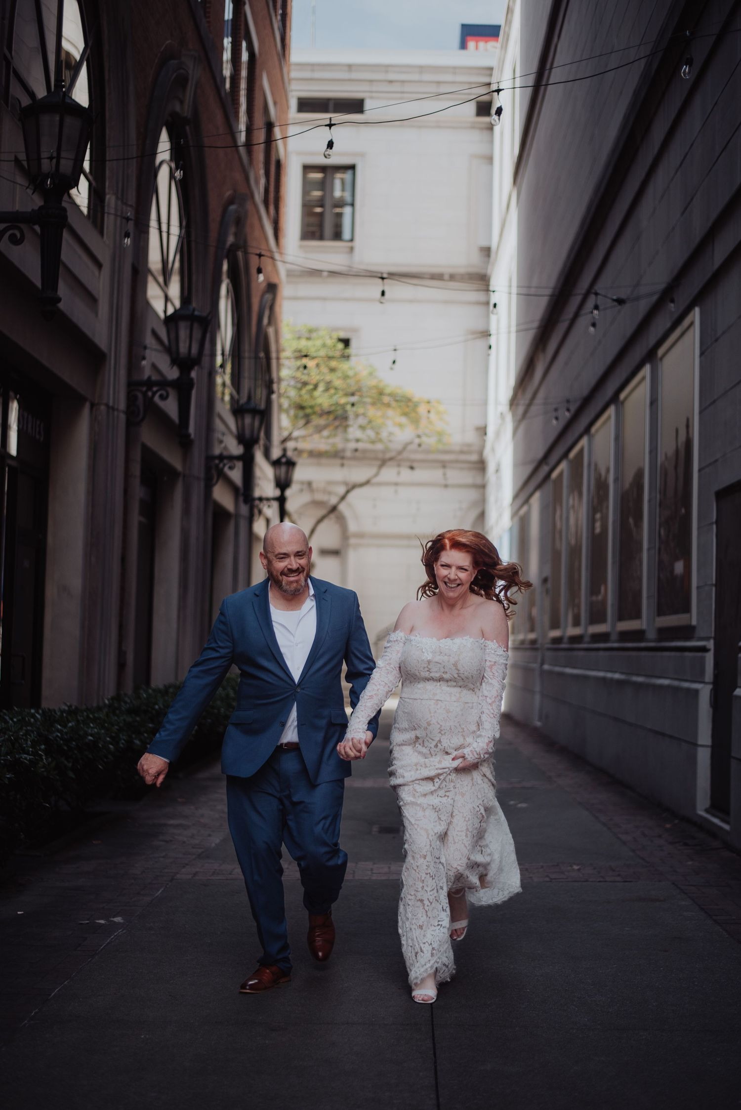 Couple running playfully down urban alley in formal attire.