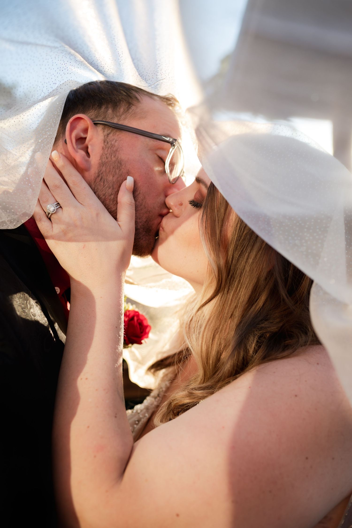 Intimate wedding moment captured under flowing white veil with sunlight streaming through.