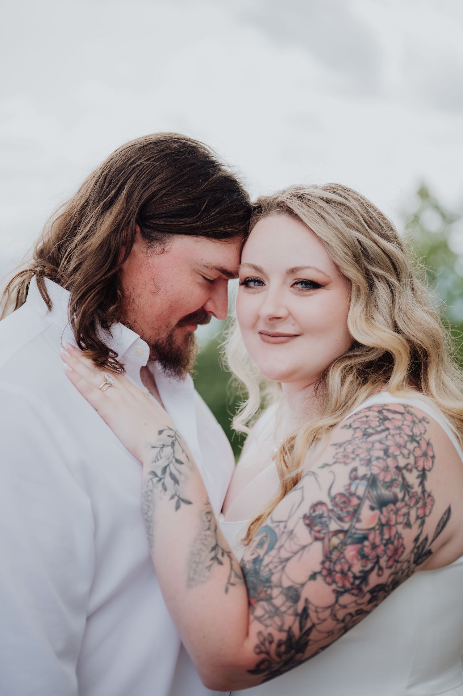 A tattooed couple in wedding attire touch foreheads tenderly, sharing an intimate romantic moment outdoors.