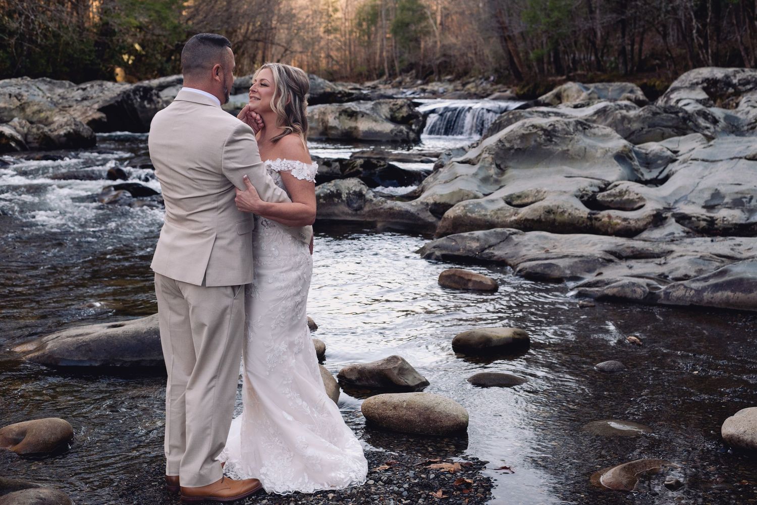 Bride and groom embracing on rocky riverbank with waterfall backdrop during outdoor wedding ceremony.