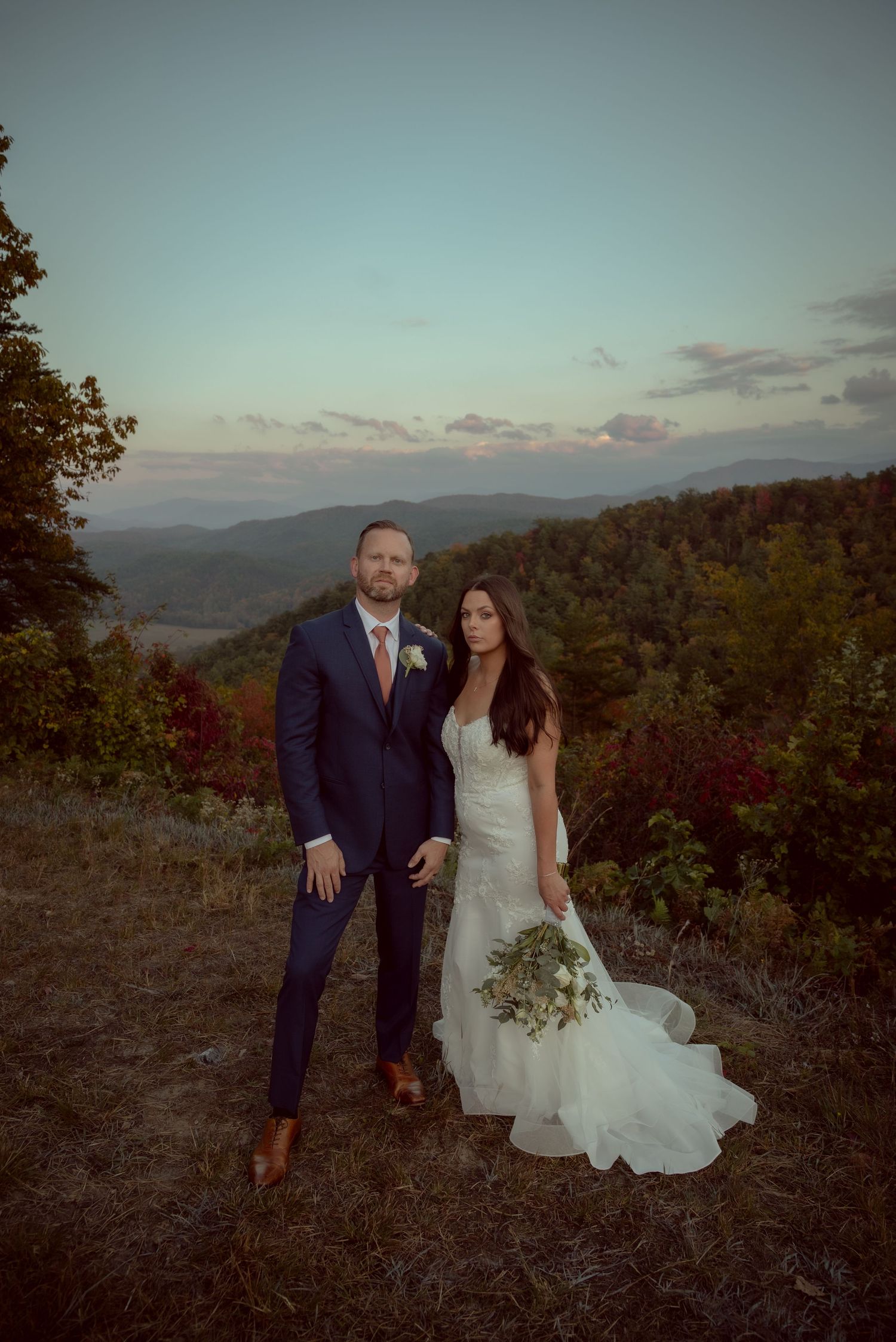 Bride and groom pose at sunset with sweeping mountain views, she in a fitted white gown, he in a navy suit.