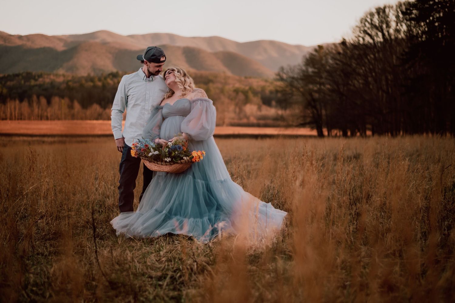 Romantic sunset photoshoot in a golden field with flowing blue dress and mountains in background.