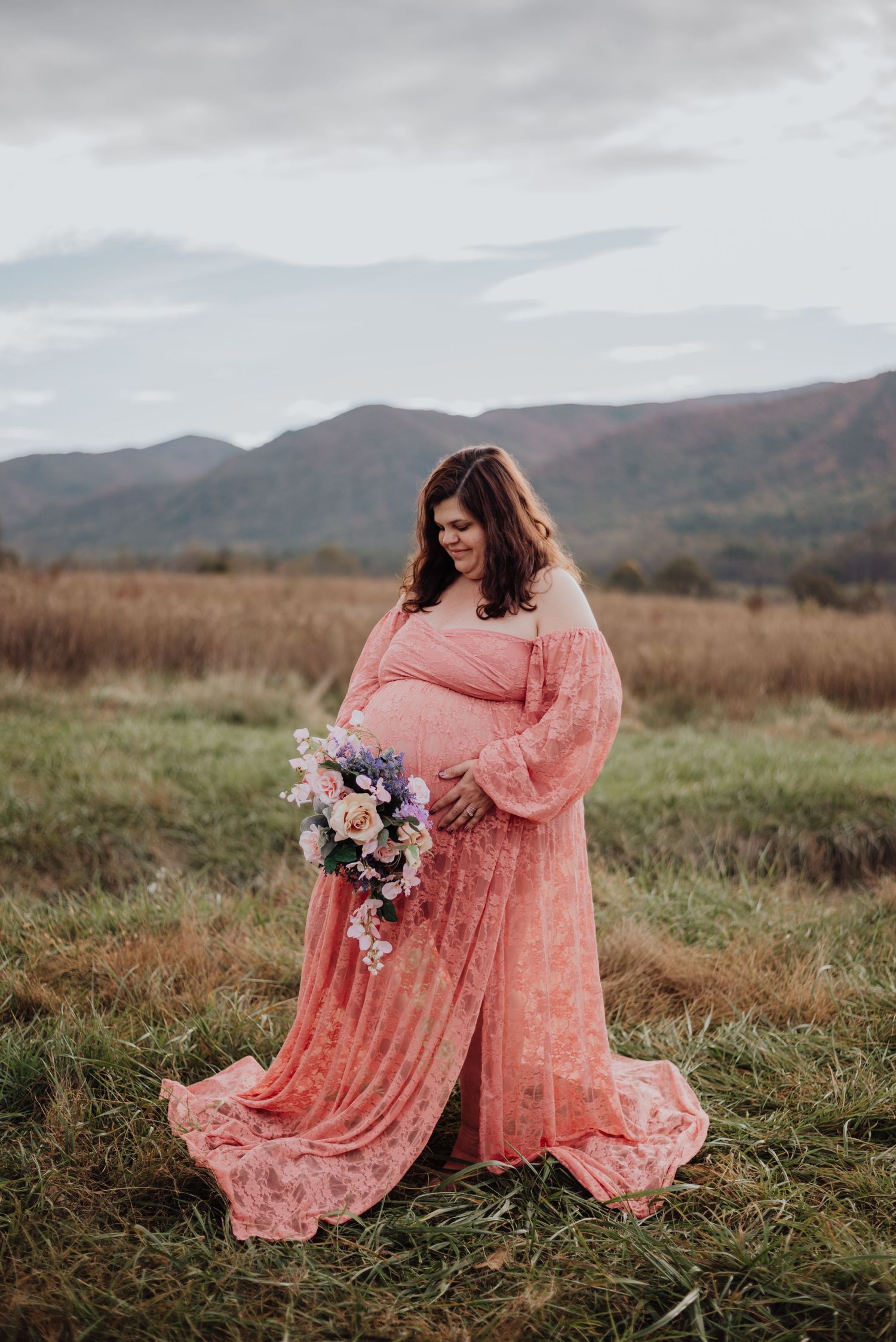 Maternity photo of woman in coral lace dress holding flowers against mountain backdrop.