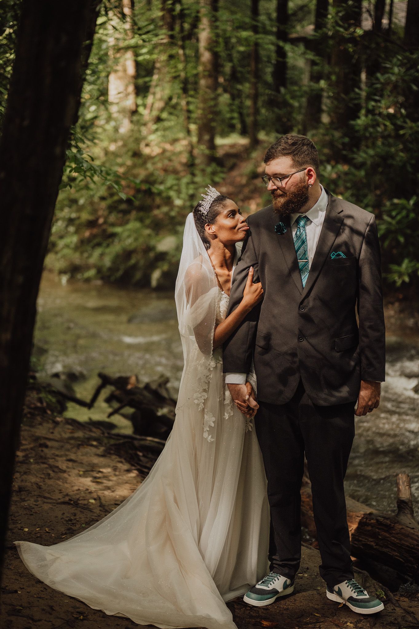 Bride and groom share a romantic moment in a lush forest setting, bride in flowing gown with veil, groom in gray suit.