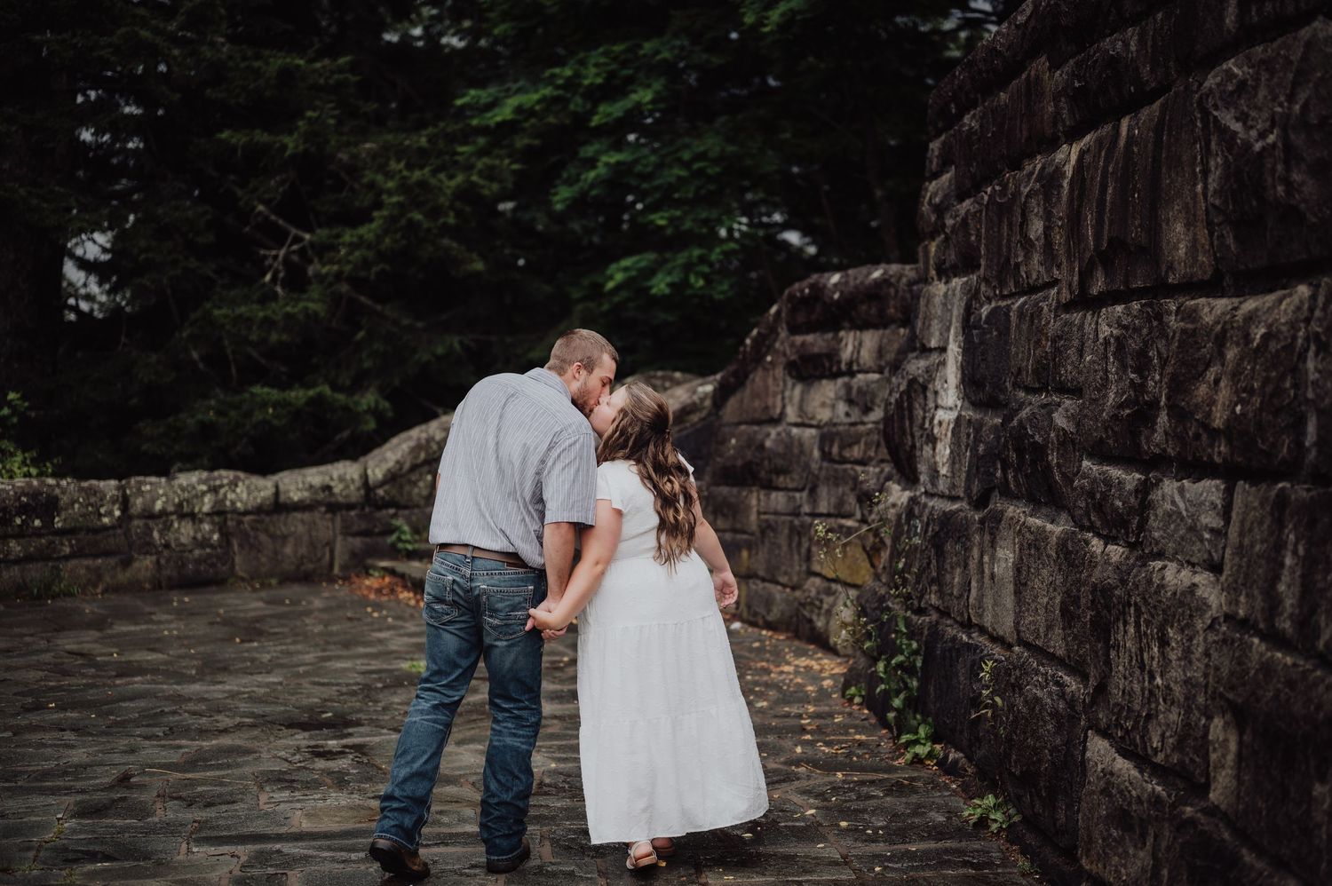 Couple sharing a tender kiss while holding hands near a stone wall outdoors.