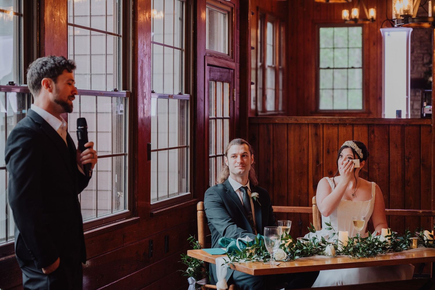 A man gives a wedding toast while the bride and groom sit at a decorated table in a wood-paneled reception hall.