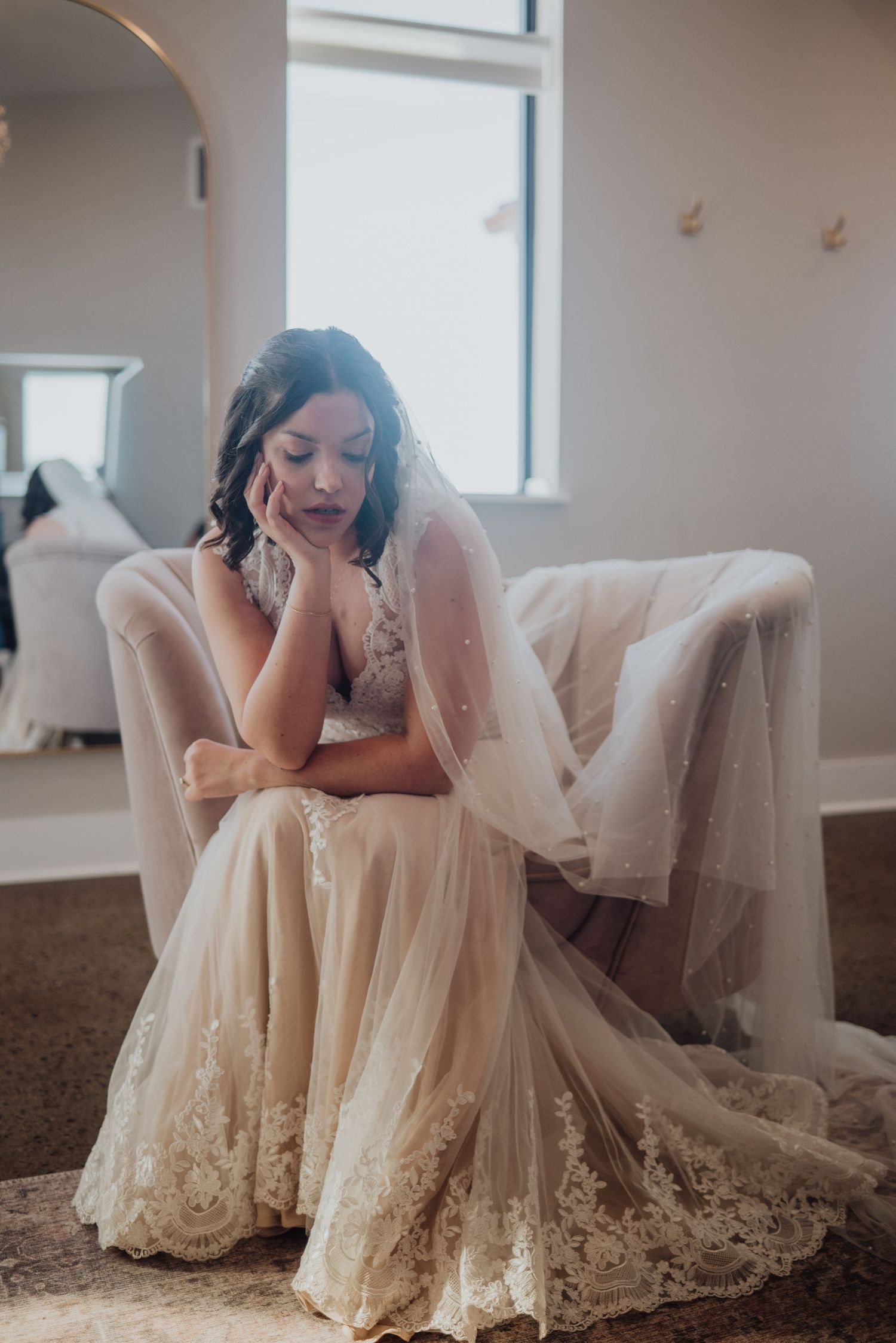 Bride in lace wedding gown and veil sits pensively on chair in bright bridal suite before ceremony.