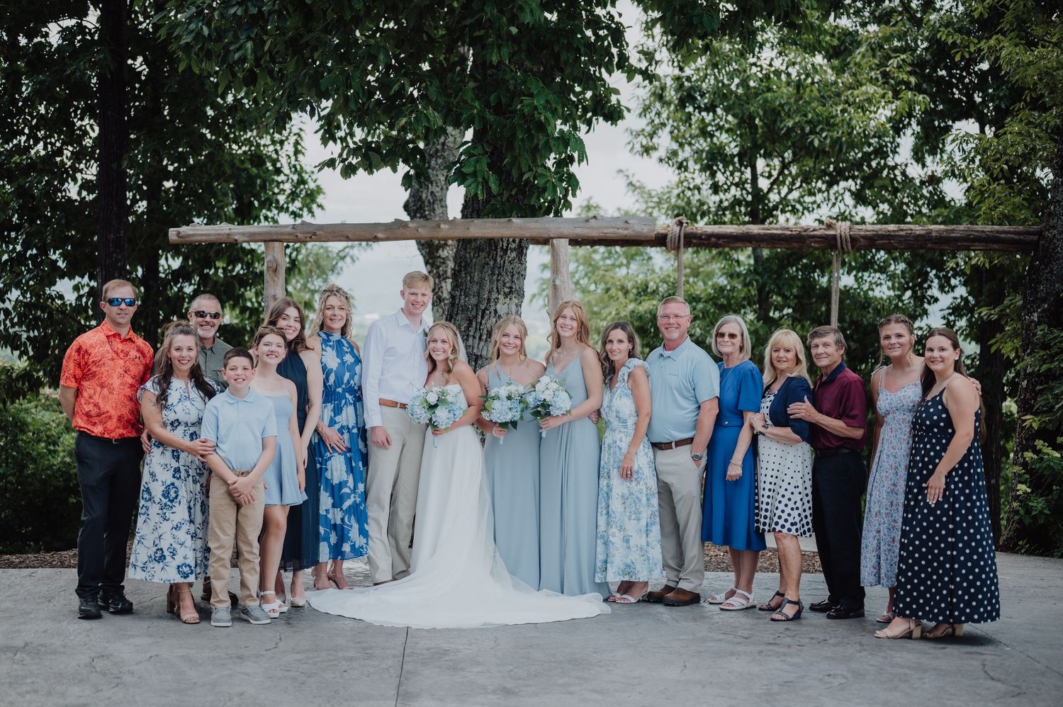 Wedding party group photo with bride and groom surrounded by family under a rustic wooden arch outdoors.