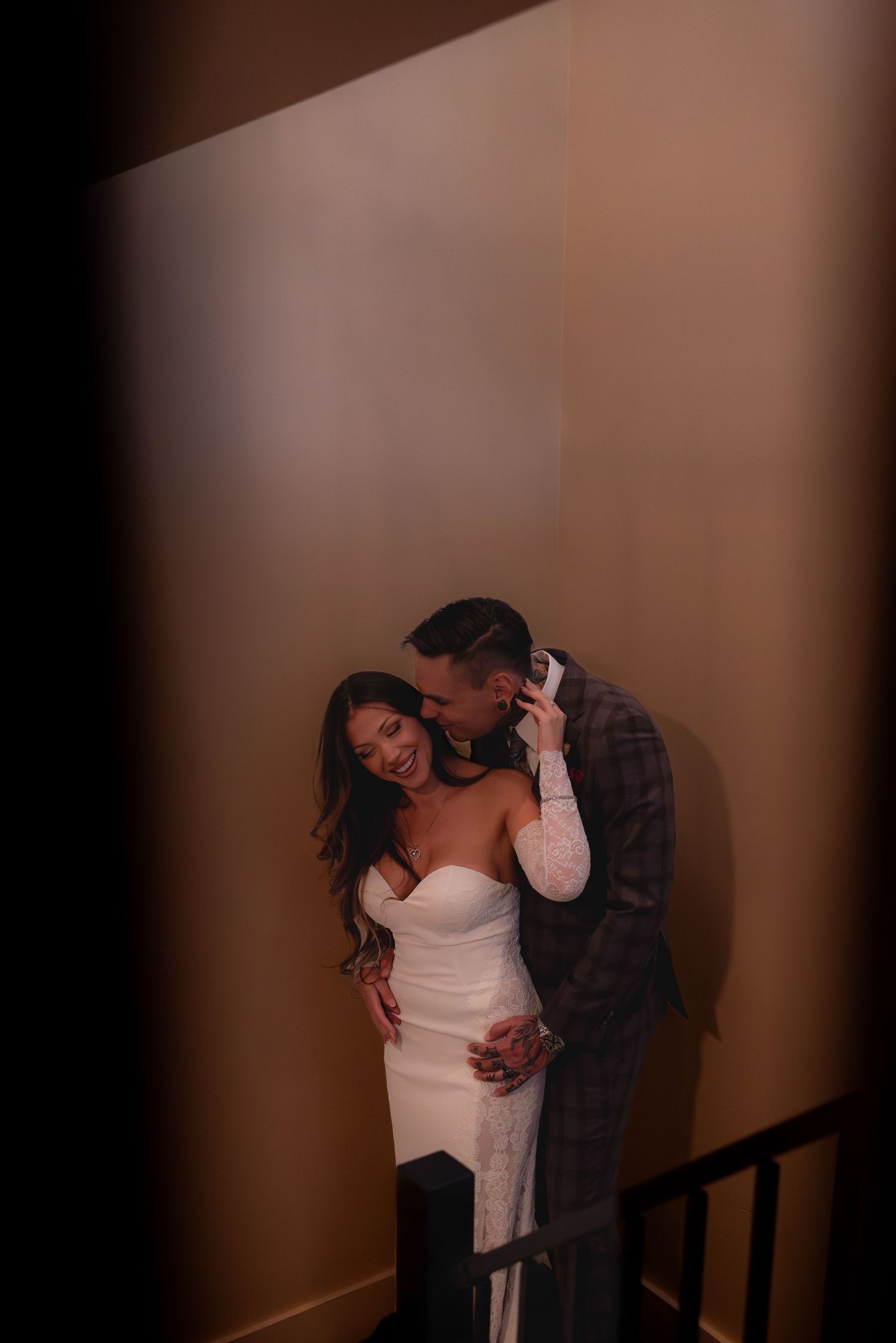 A bride in a white strapless gown and groom in plaid suit share a romantic embrace on a dimly lit staircase.