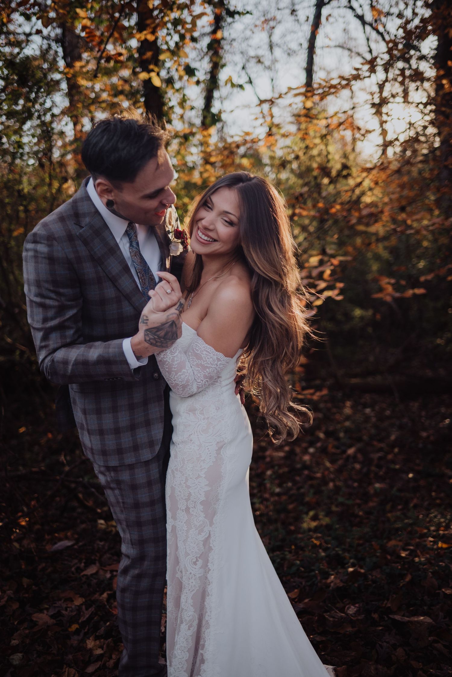 Bride and groom embracing in autumn forest, she smiles in lace gown, he wears plaid suit, golden foliage backdrop.
