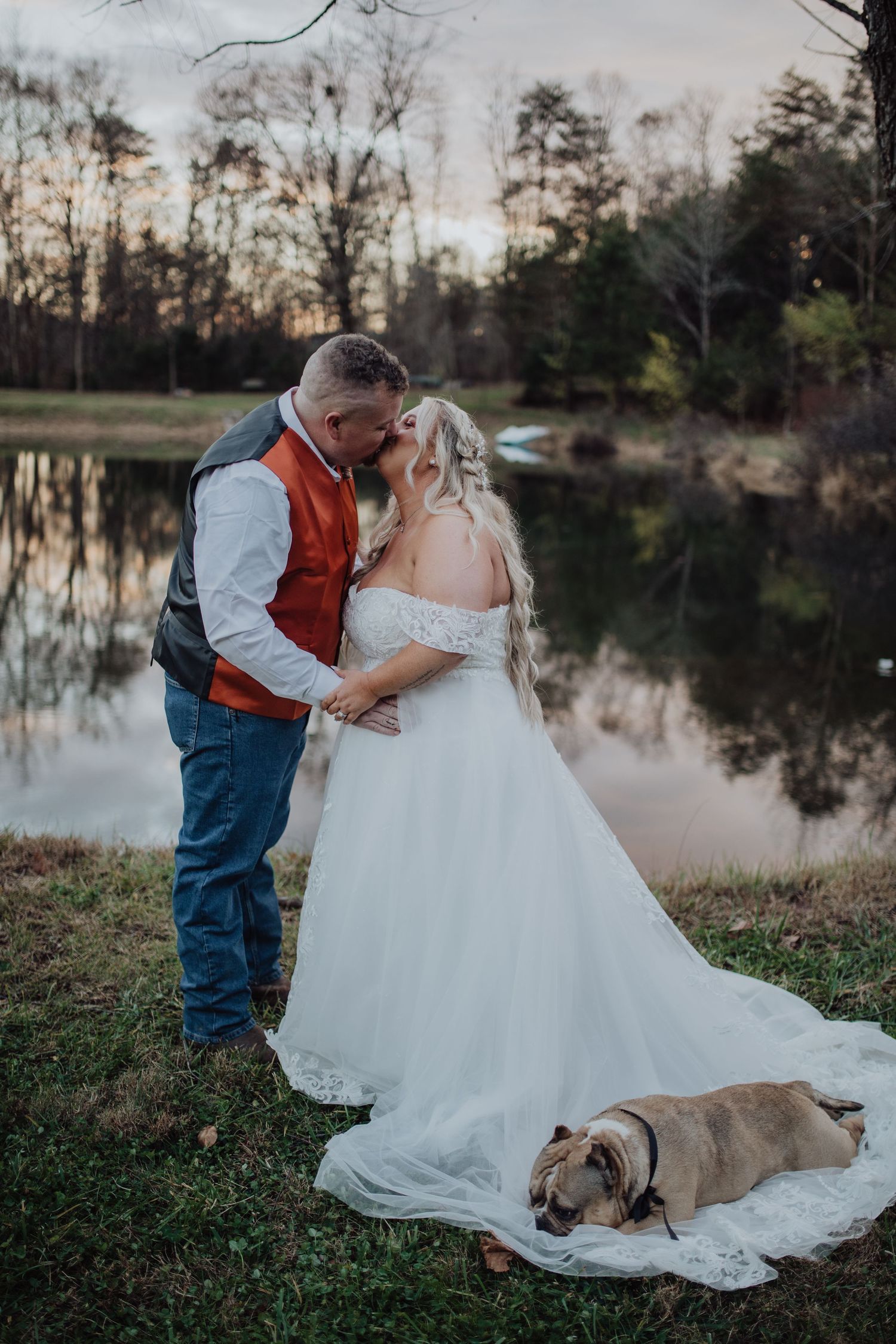 Bride and groom share a kiss by a reflective pond at sunset, with their bulldog resting on the wedding dress train.