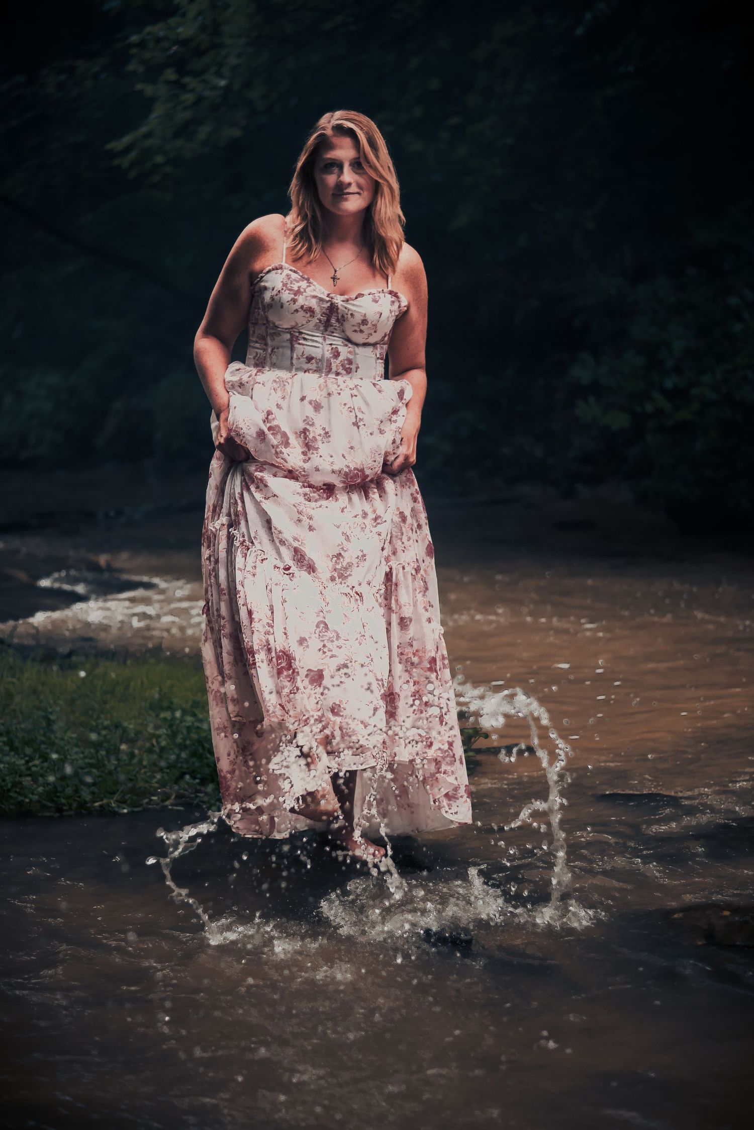 Person in vintage floral dress splashing in shallow water during moody photoshoot.