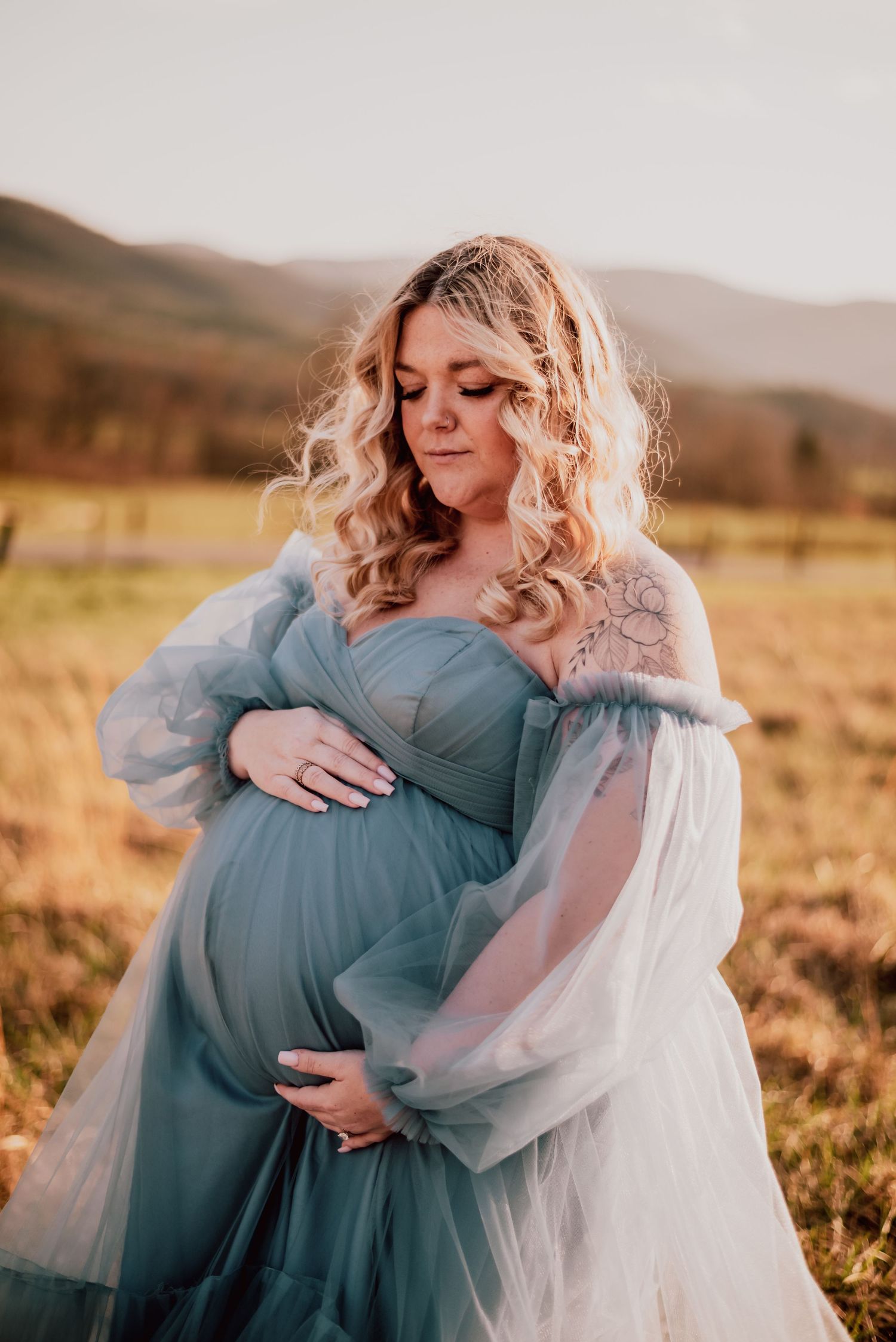 Expectant mother in flowing blue tulle gown poses in golden field with mountains behind.