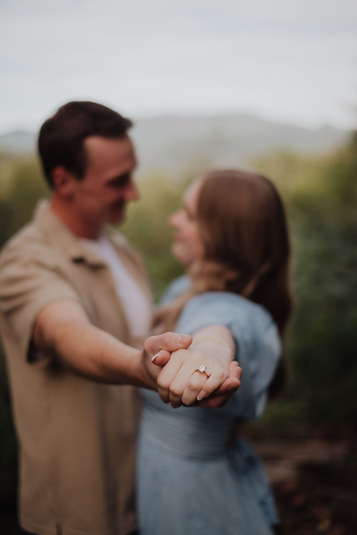 A close-up engagement photo showing clasped hands with a ring in soft focus against a natural outdoor background.