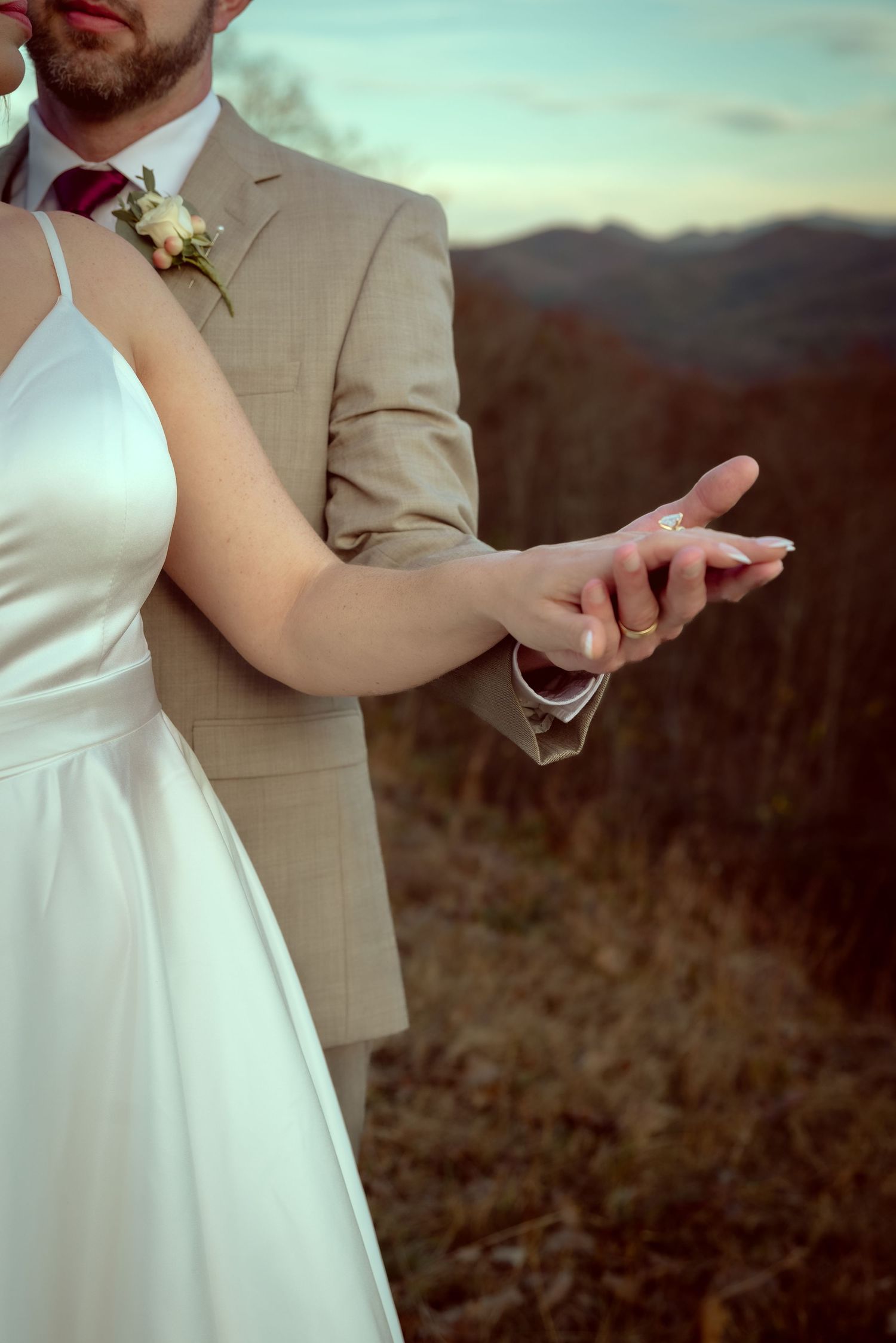Close-up details of wedding couple's hands and formal attire against mountain backdrop.