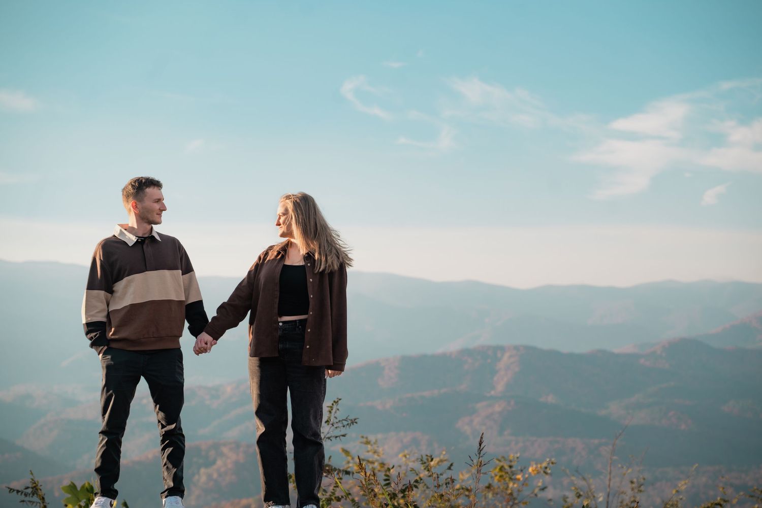 Couple holding hands while standing on mountain overlook against blue sky and distant peaks.