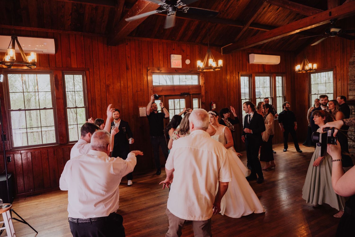 Guests dancing joyfully in a rustic wood-paneled wedding reception hall with warm chandelier lighting.