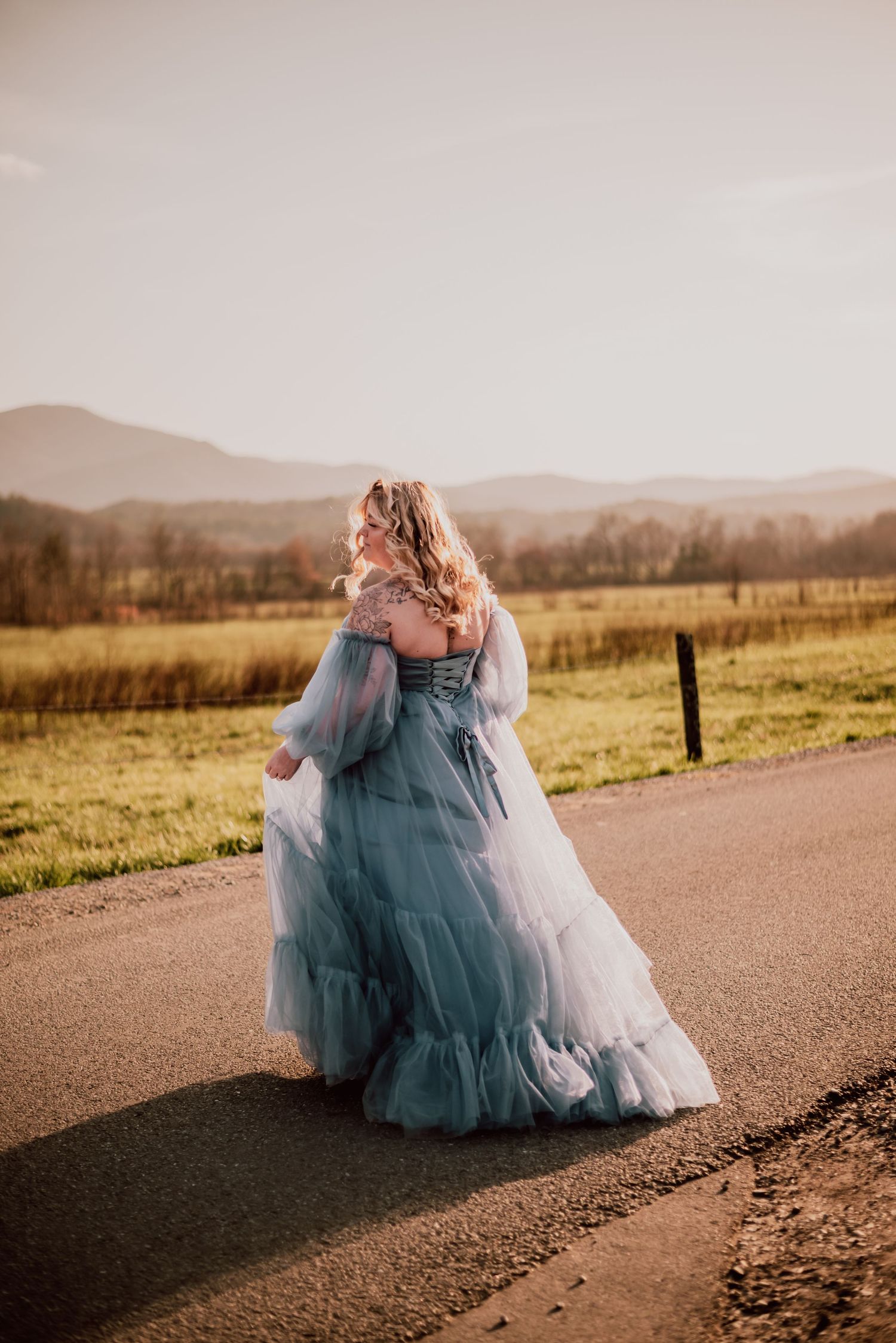 Woman in ethereal blue maternity gown walks along country road with mountains in background.