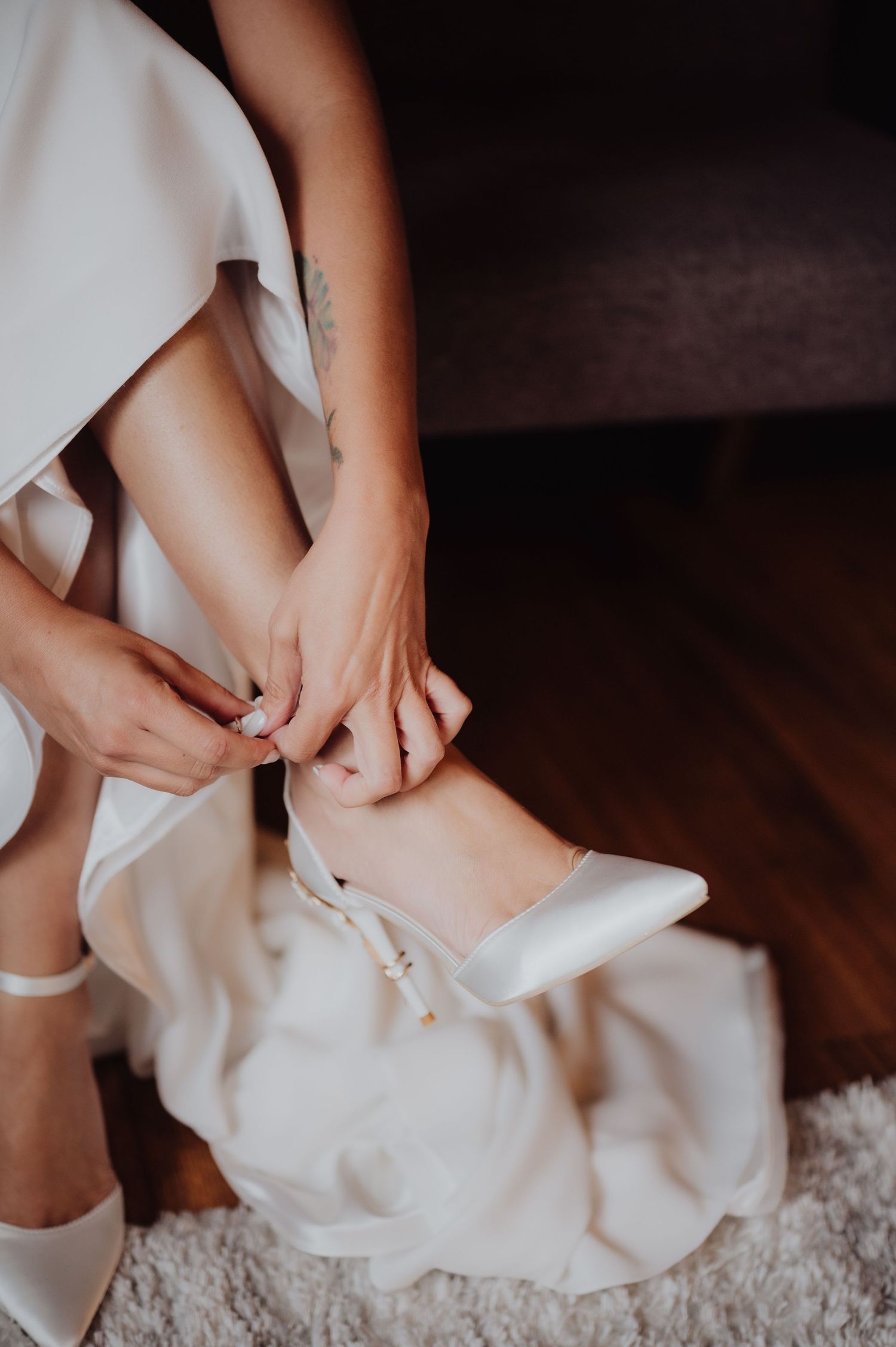 Bride fastening white satin heels while wearing wedding dress, seated on floor with soft rug.