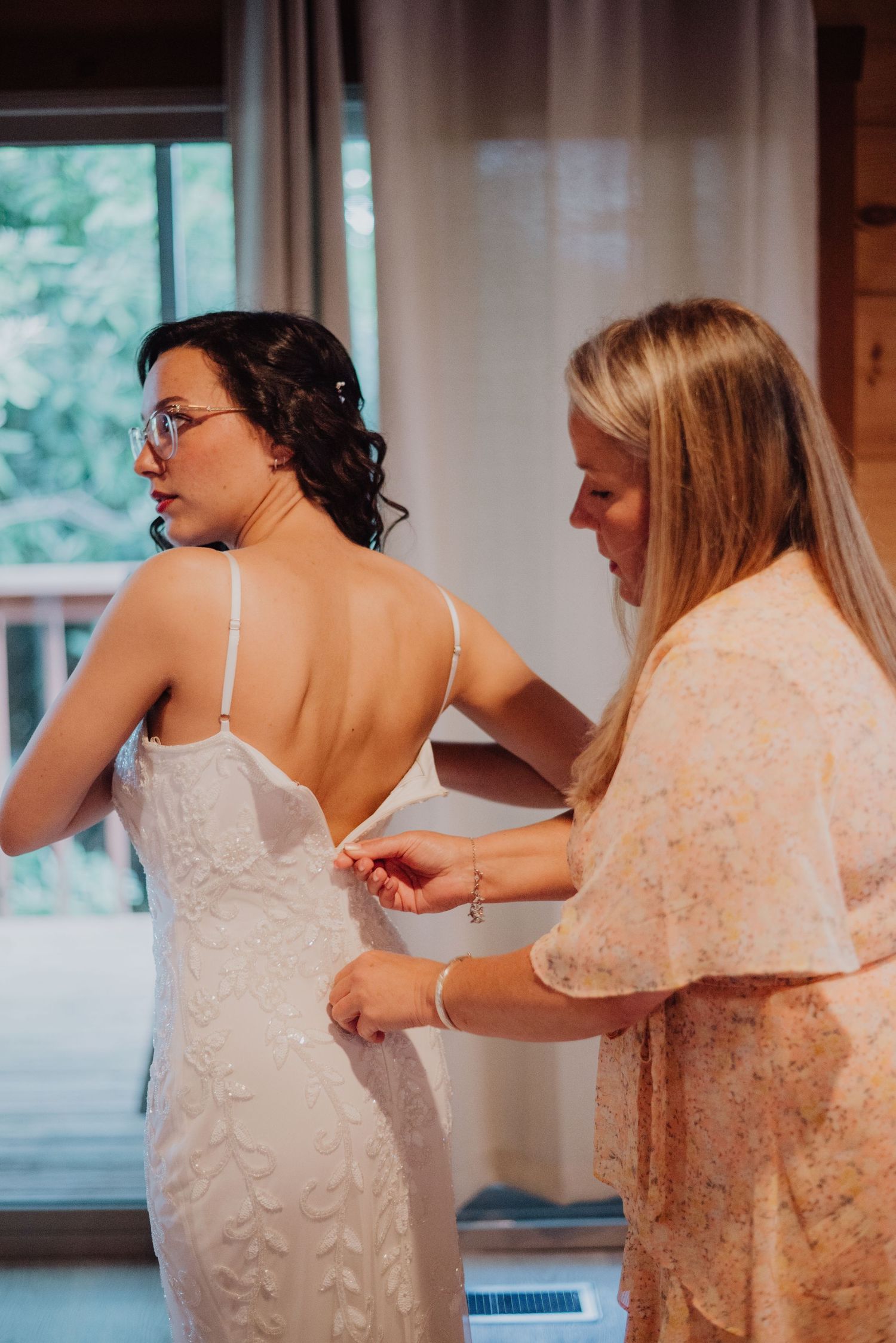 A bridesmaid helps a bride in a white lace backless gown get dressed before her wedding ceremony.