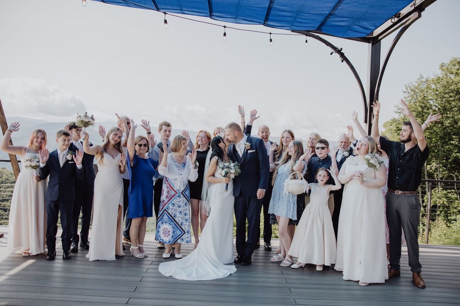 Wedding party celebrating outdoors under a pergola, bride and groom kissing while guests cheer around them.