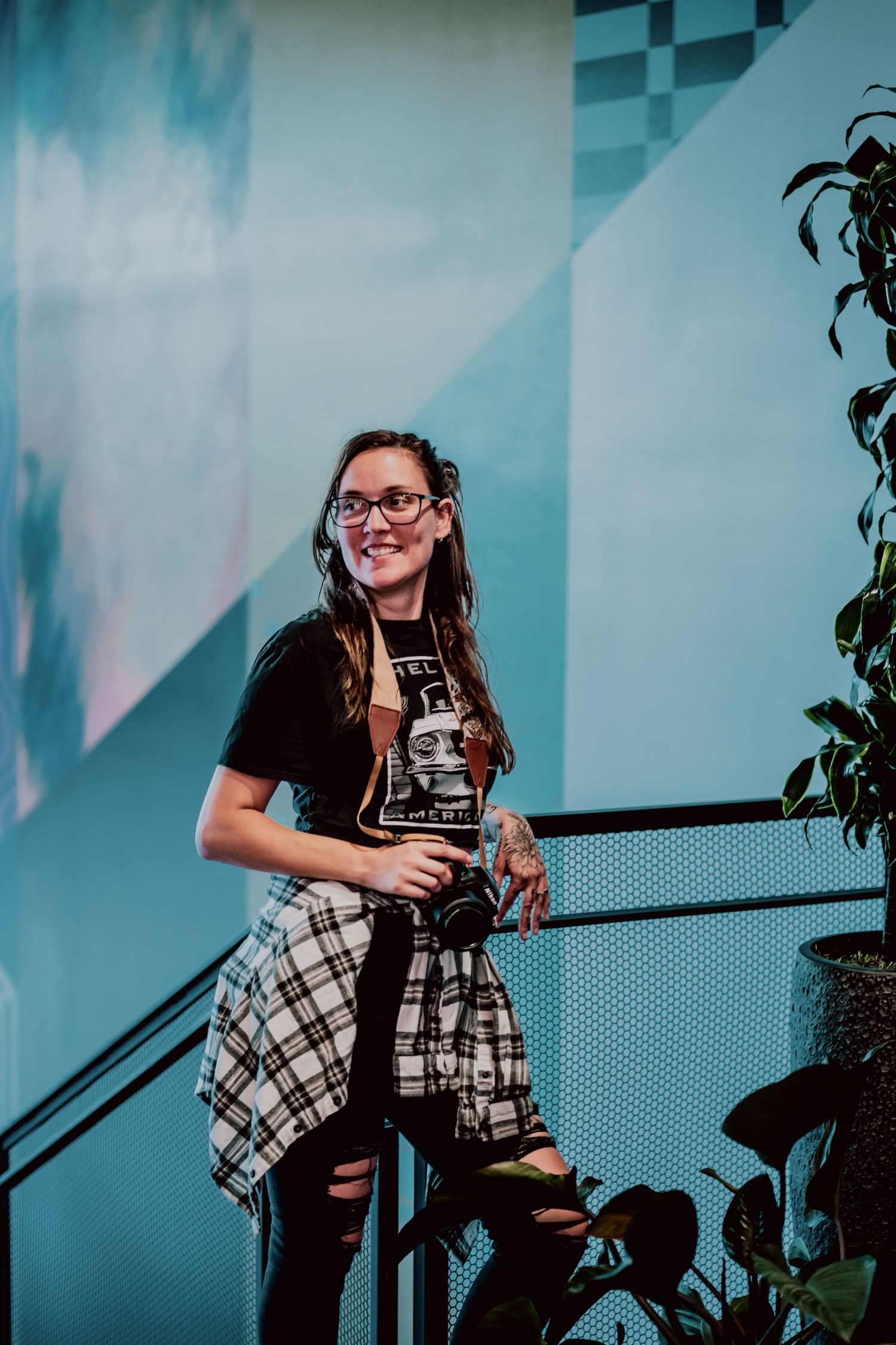 A stylish person in glasses and casual attire poses against a blue geometric wall with plants.