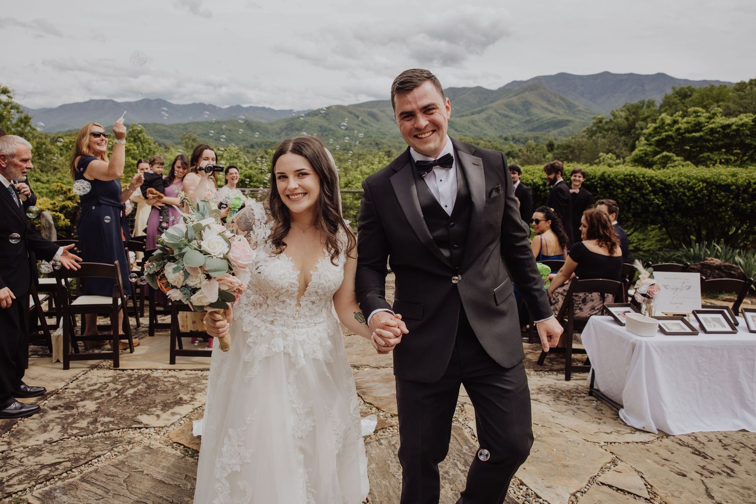 Newlywed couple holding hands and smiling after outdoor wedding ceremony with lush mountain views.