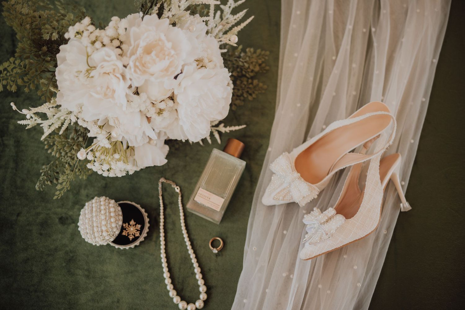 Elegant wedding flat lay featuring white peony bouquet, pearl necklace, heels, perfume, and bridal accessories on green velvet.
