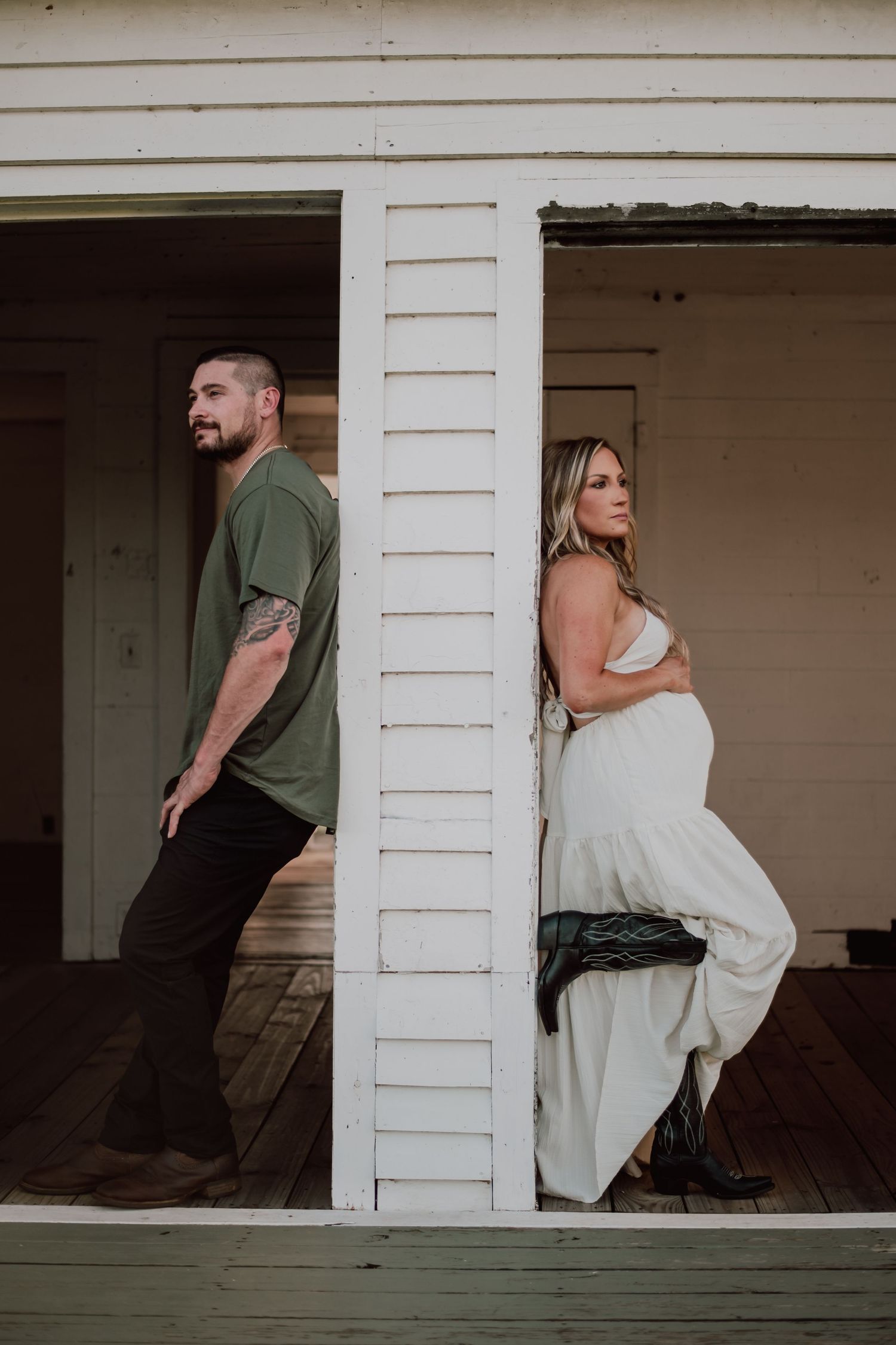 A couple poses back-to-back on a rustic white porch for a maternity photo.