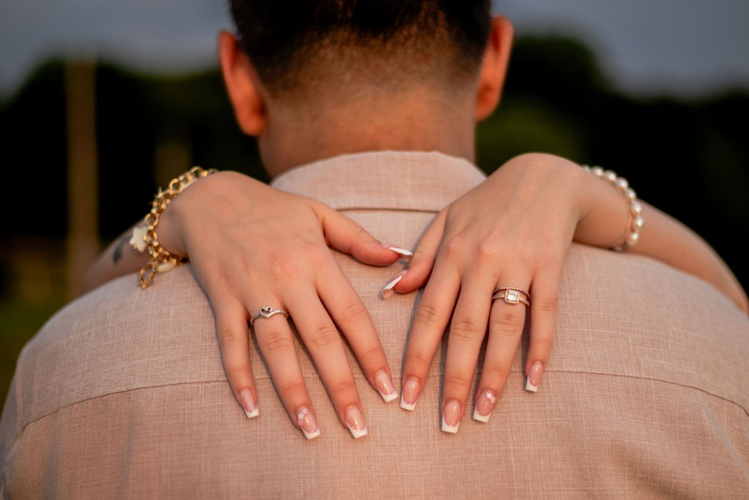 Close-up of hands with rings embracing from behind against a beige suit jacket.