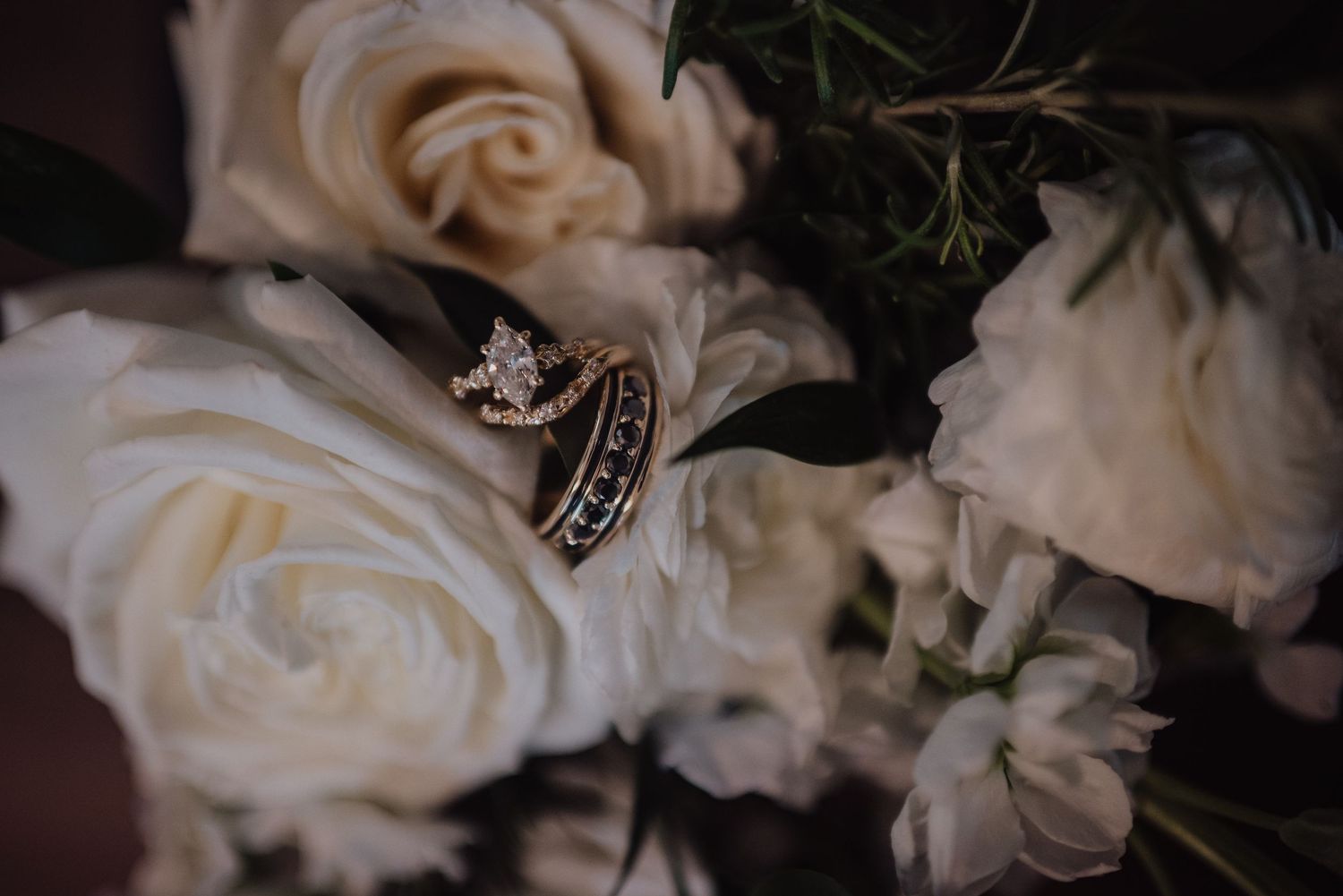 Gold wedding and engagement rings with diamond resting on white roses in a bridal bouquet.
