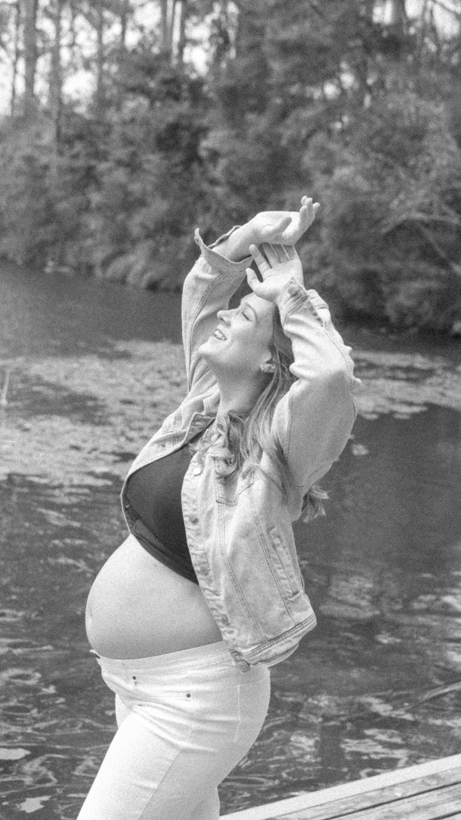 Black and white maternity photo of a pregnant woman in a denim jacket and white pants smiling with arms raised beside a calm lake.