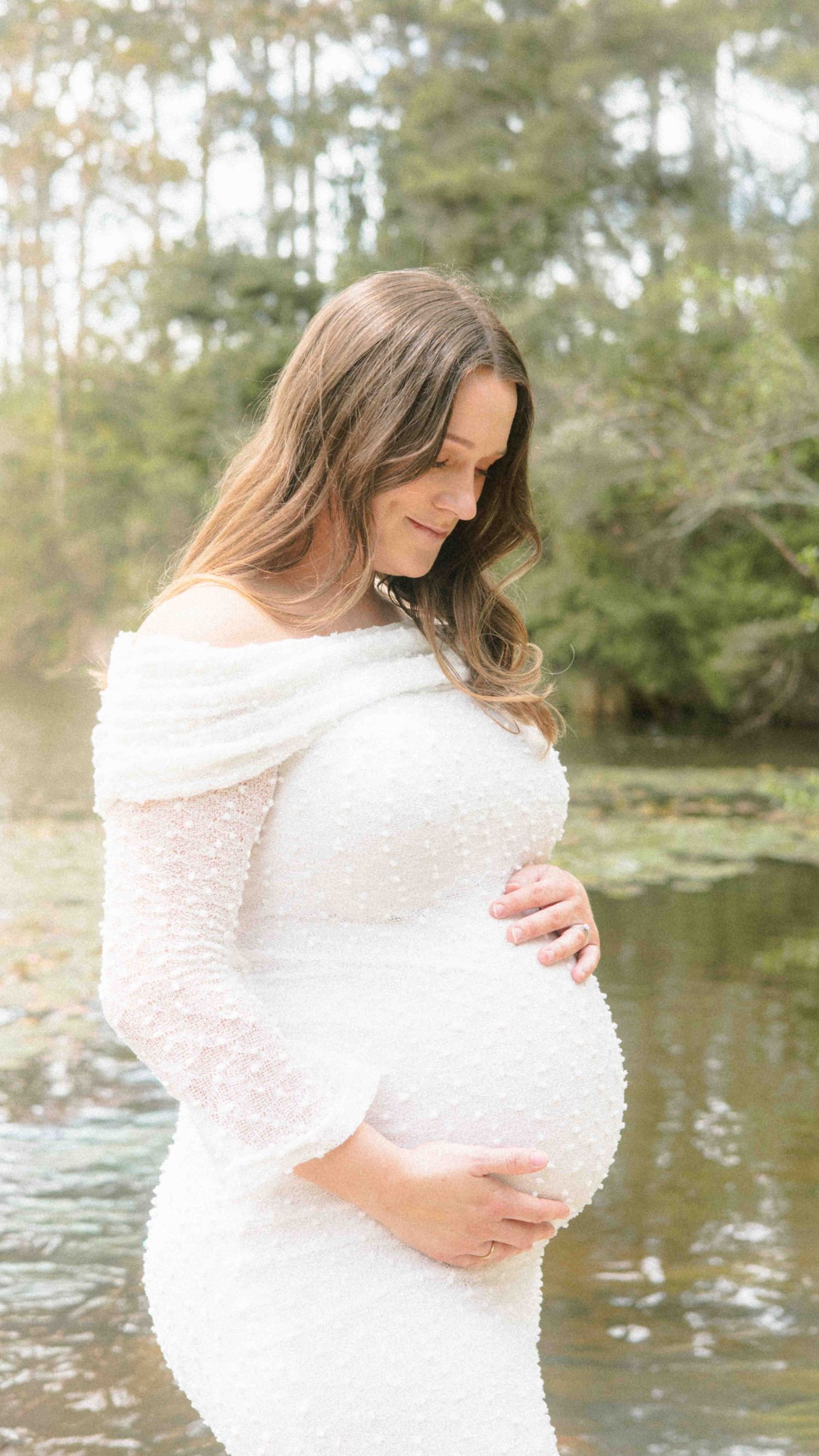 Pregnant woman in a white off-the-shoulder maternity gown cradling her baby bump beside a calm lake with trees in the background.