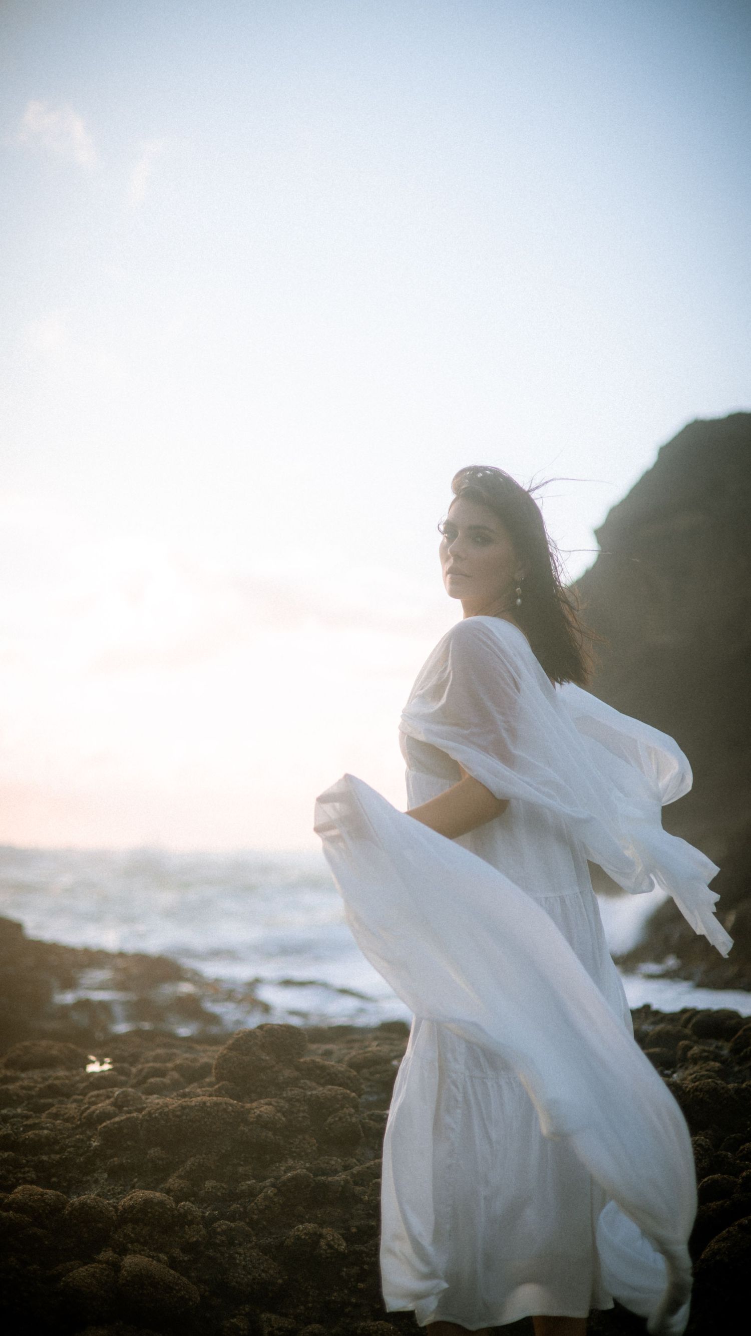 Dynamic movement captured in white dress against moody coastal landscape at sunset.