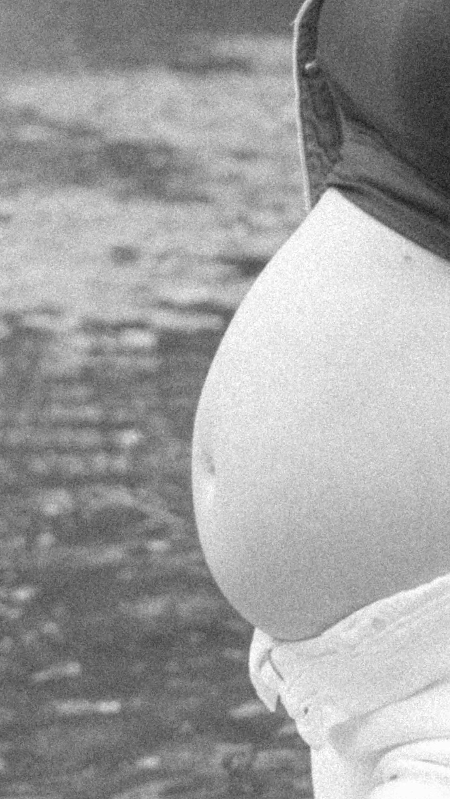 Black and white close-up of a pregnant woman's bare belly in profile, wearing white pants and a dark top outdoors.