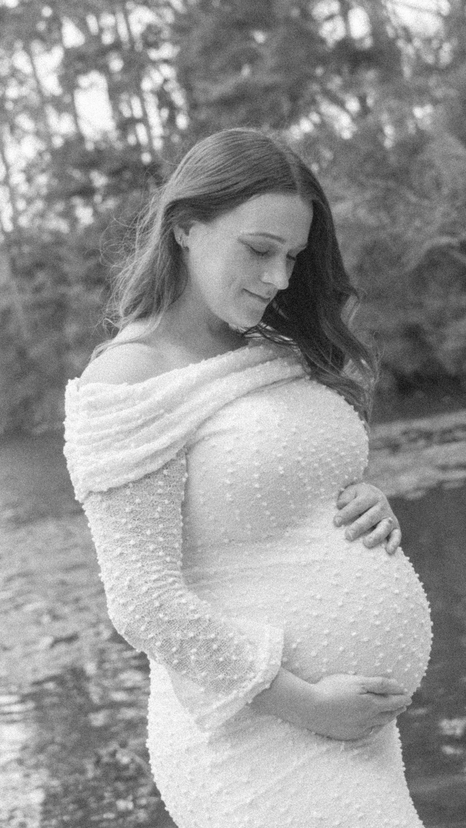 Black and white maternity portrait of a pregnant woman in an off-shoulder lace gown cradling her baby bump beside a tranquil river.