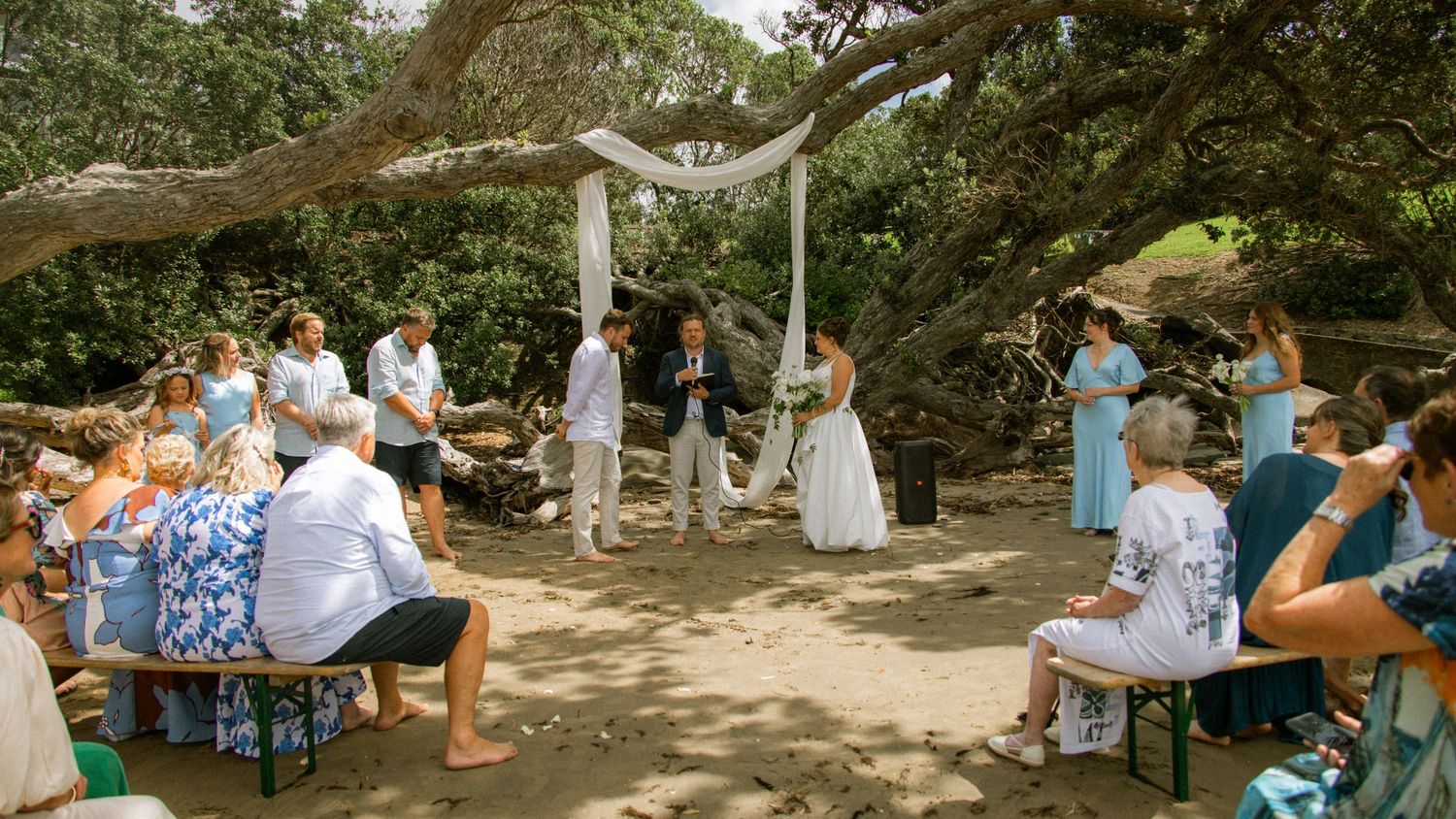 Outdoor wedding ceremony under a large tree with couple at altar, guests seated on chairs in a natural garden setting.