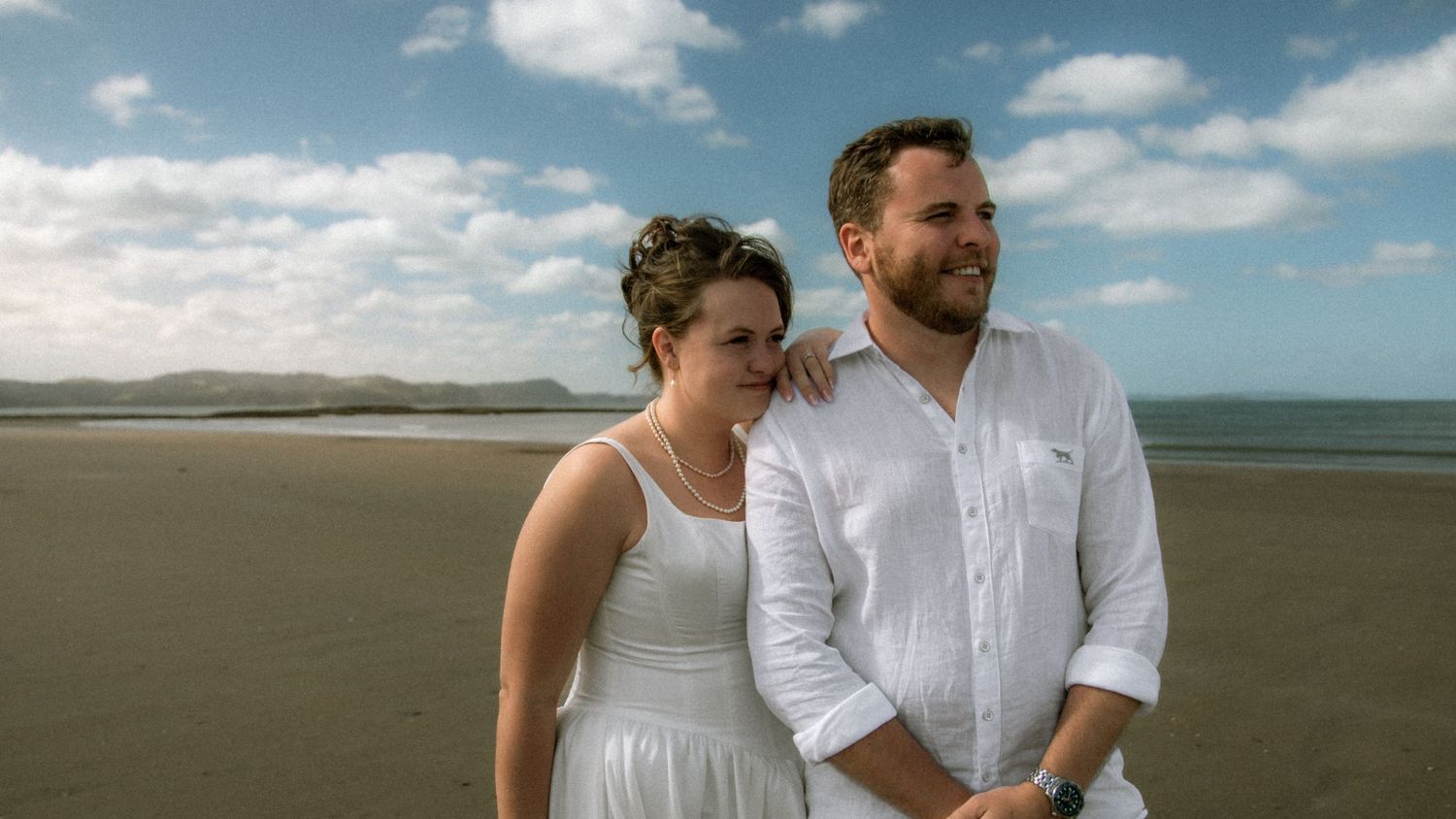 A couple in white outfits walks along a scenic beach shoreline with dramatic cloudy skies above.