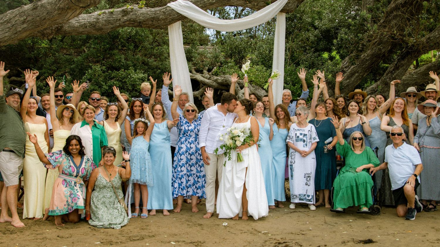 Joyful wedding group photo outdoors under a white arch with bridesmaids in blue dresses celebrating with the couple.
