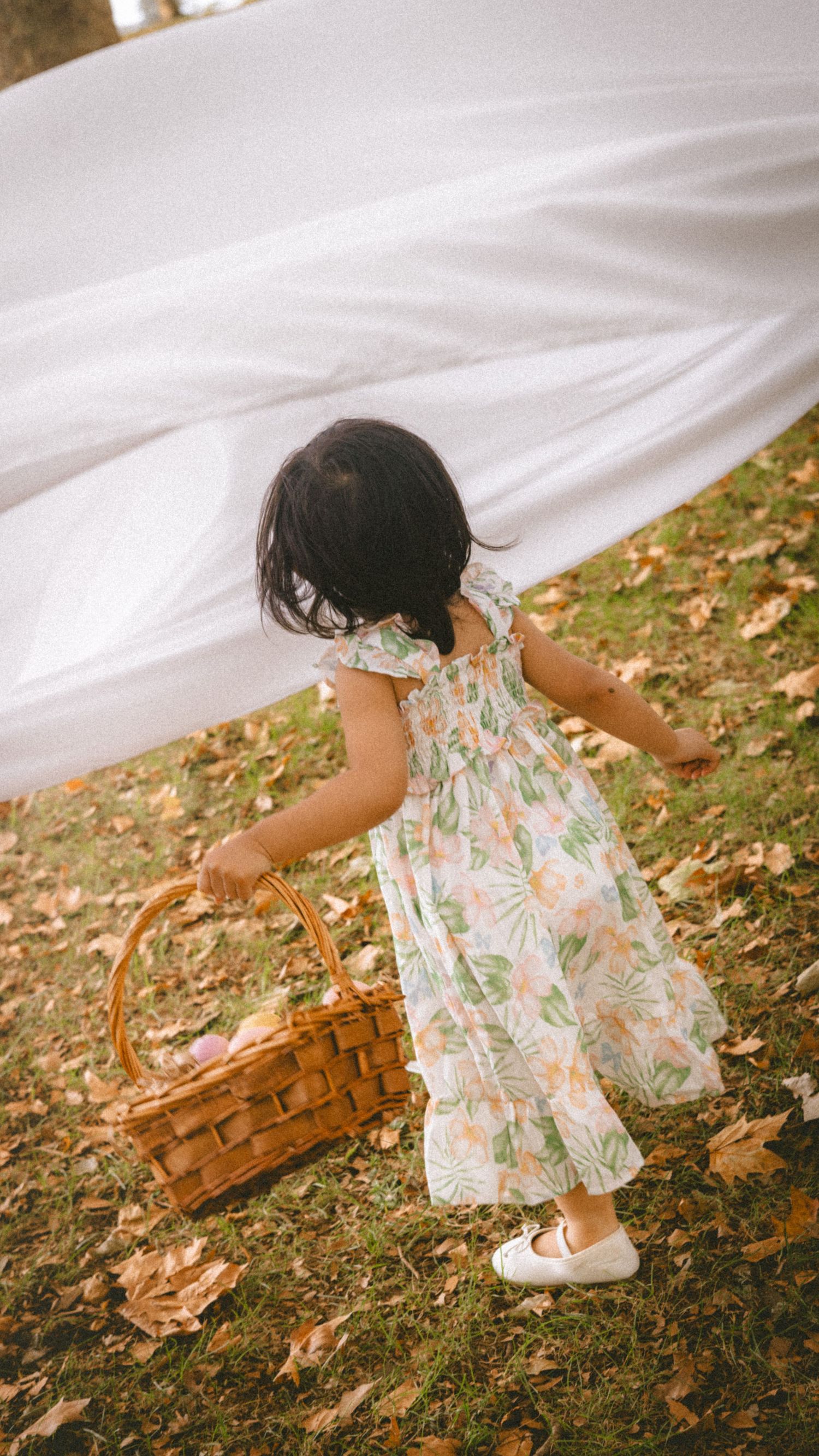 Young girl in floral dress carrying wicker basket on grass with autumn leaves and white fabric backdrop.