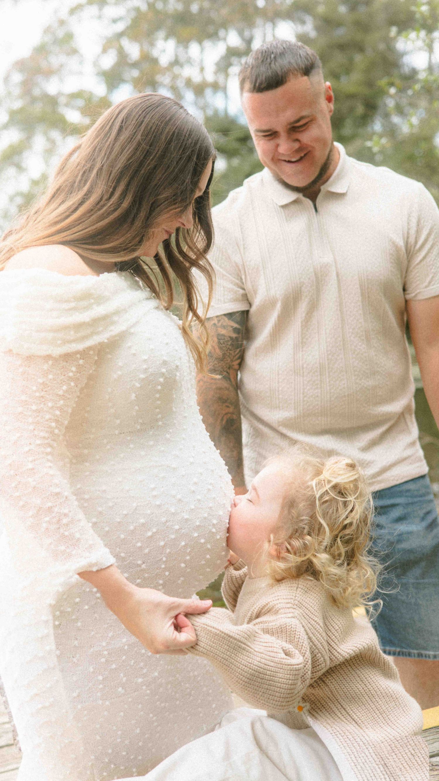 A toddler with curly blonde hair kisses the belly of a pregnant woman in a white dress while a smiling tattooed man looks on outdoors.