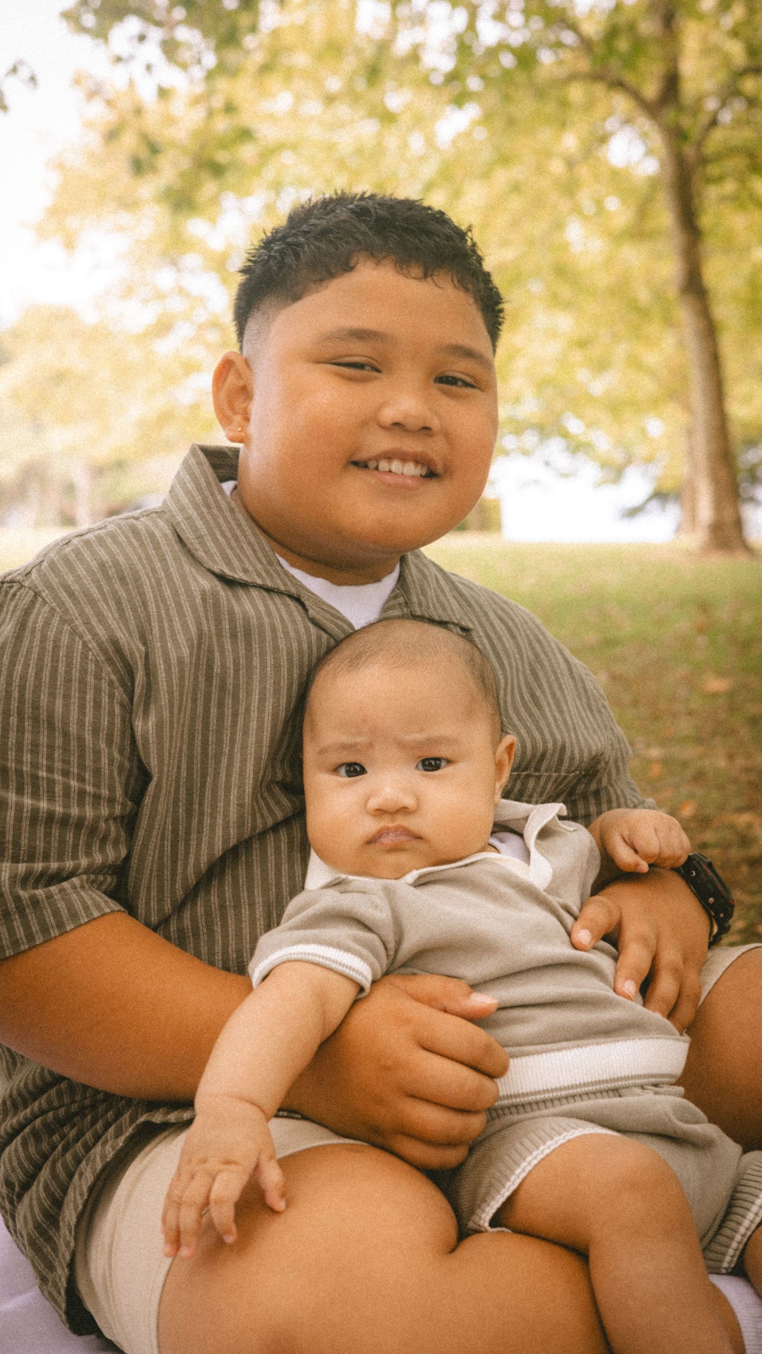 A smiling boy holds a baby in his lap outdoors in a park with autumn trees in the background.