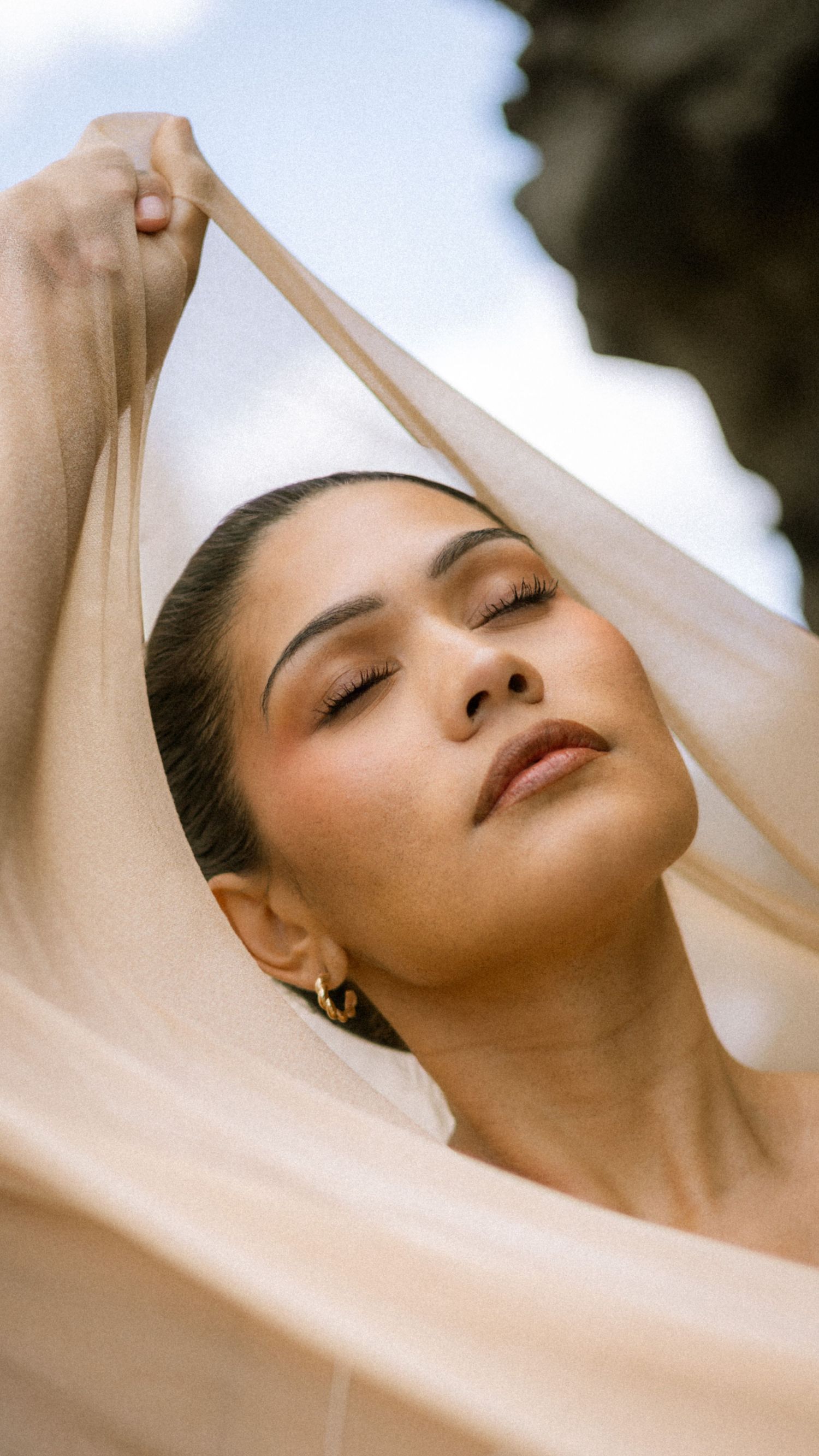 Artistic fashion portrait showing a person with closed eyes draped in flowing white fabric against a light background.