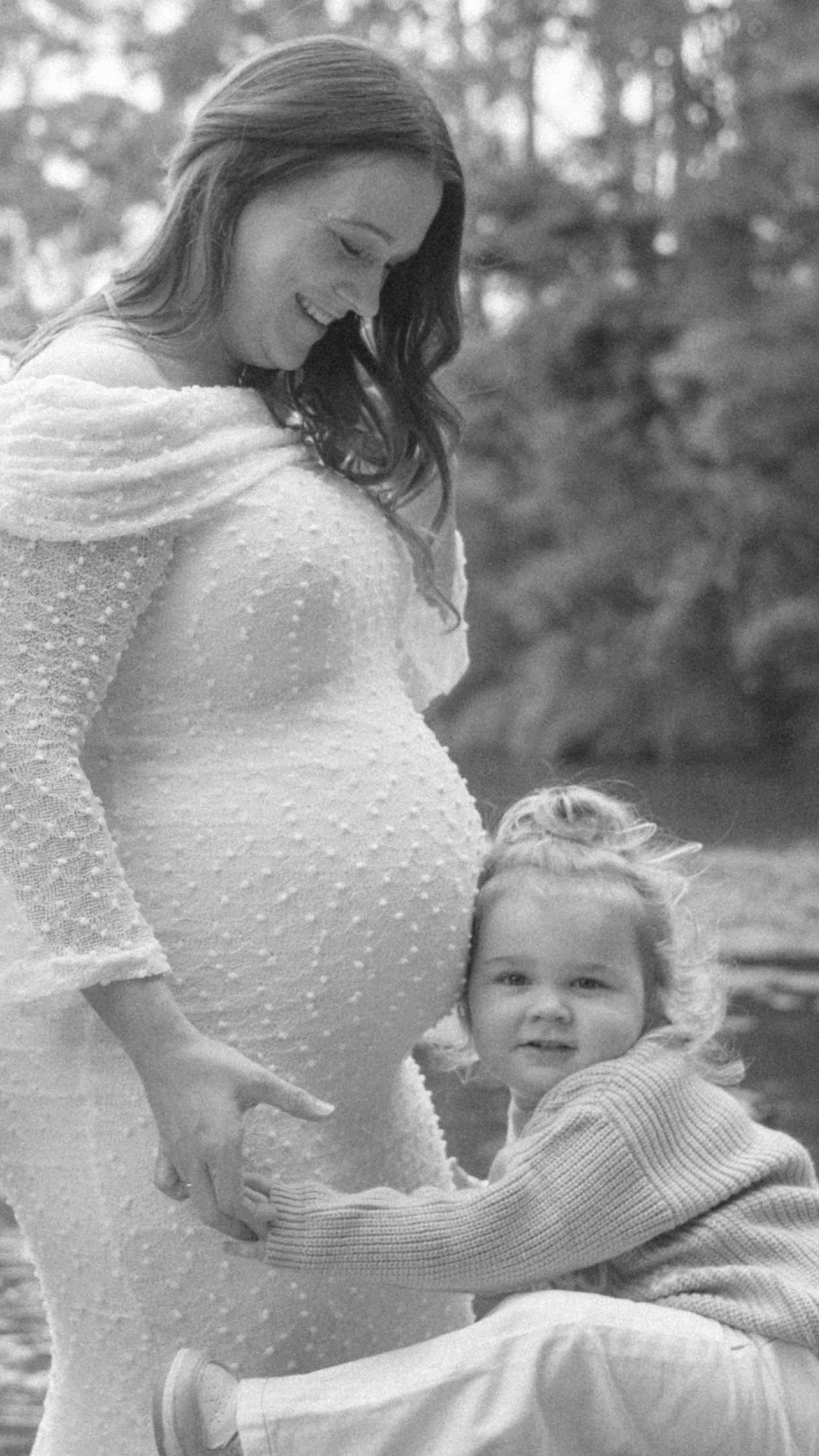 Black and white maternity photo of a smiling pregnant woman in a textured off-shoulder gown with a toddler hugging her belly outdoors.
